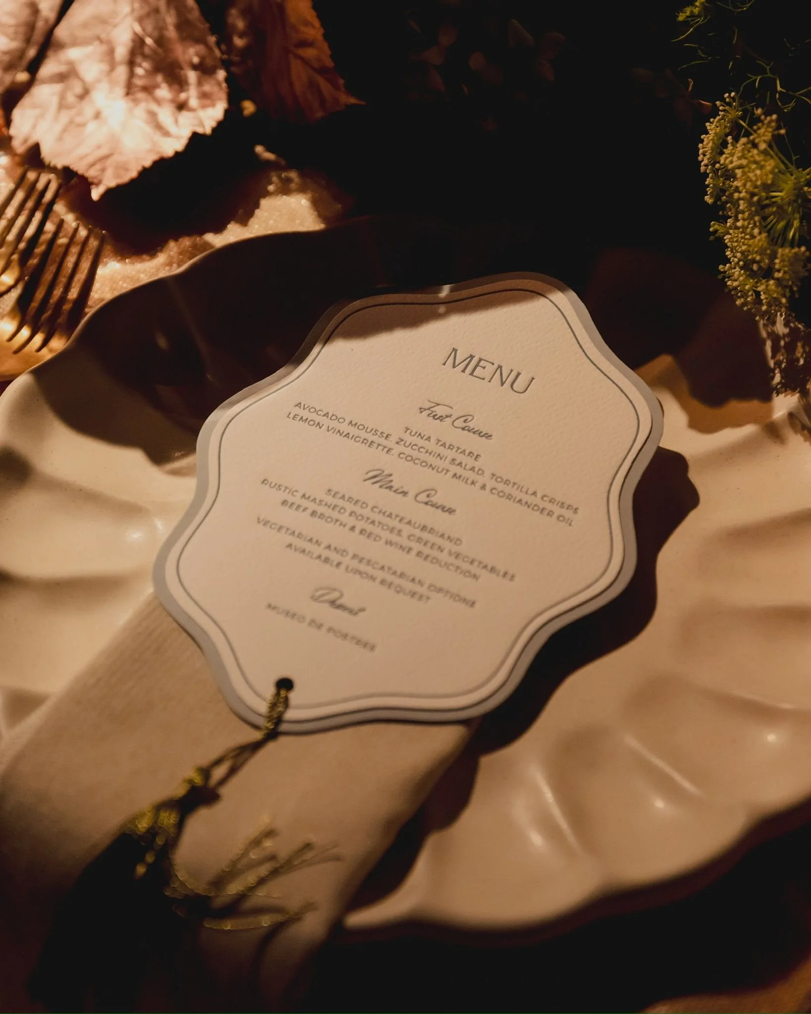 Close-up of a menu card on a table surrounded by decorative foliage, with a fork resting nearby, in warm lighting.