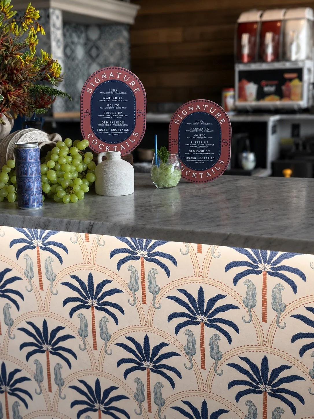 Close-up of a bar counter with a bunch of green grapes, a small white vase, a cocktail glass with a straw and mint leaves, and two pink cocktail menus listing drinks such as Luna, Margarita, Mojito, Puffer Up, Old Fashion, and Frozen Cocktails. In the background, a coffee machine and a patterned wall are visible.