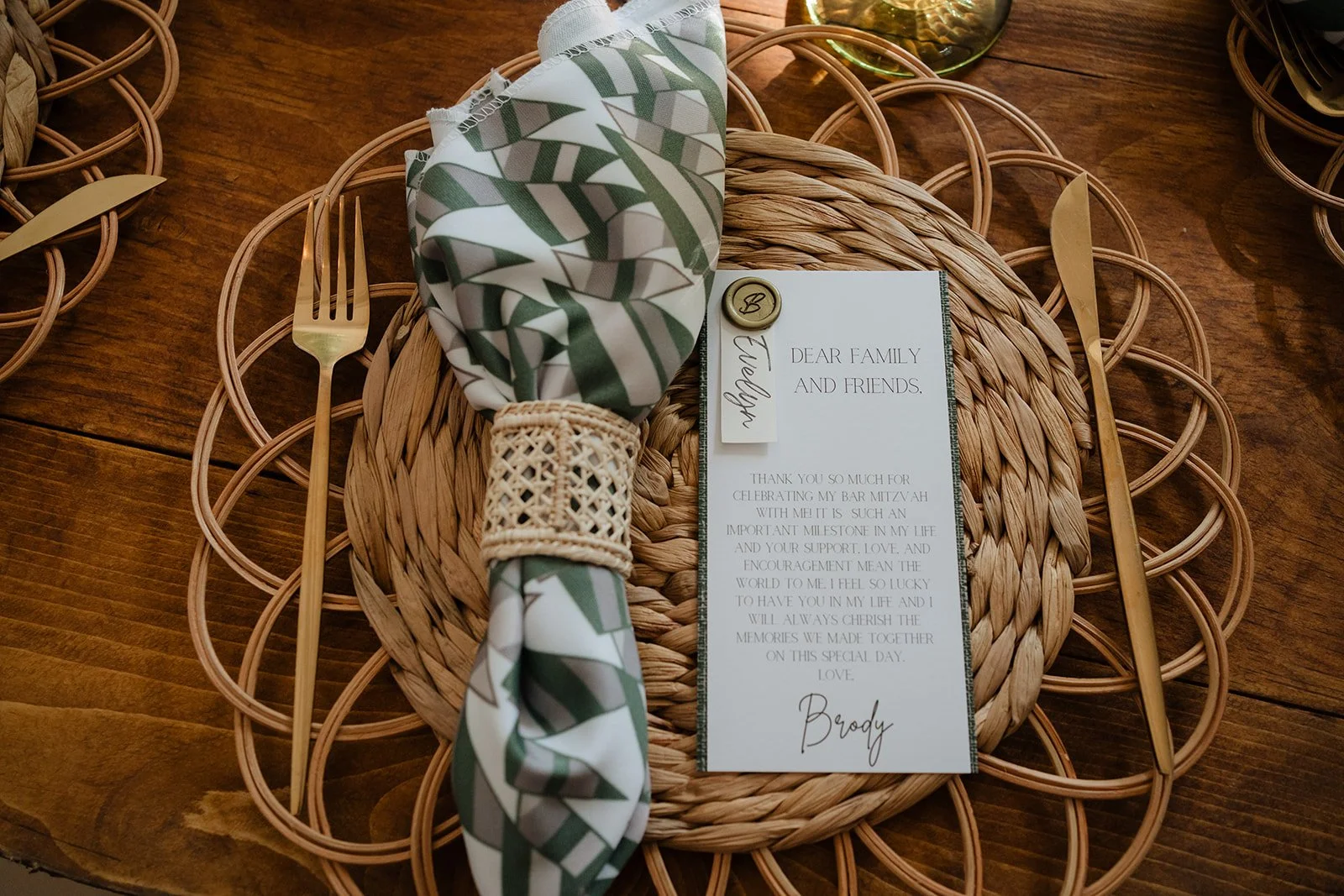 A woven placemat with gold-colored cutlery on a wooden table, a cloth napkin with green and white patterns, a small card with a message, and a decorative button.