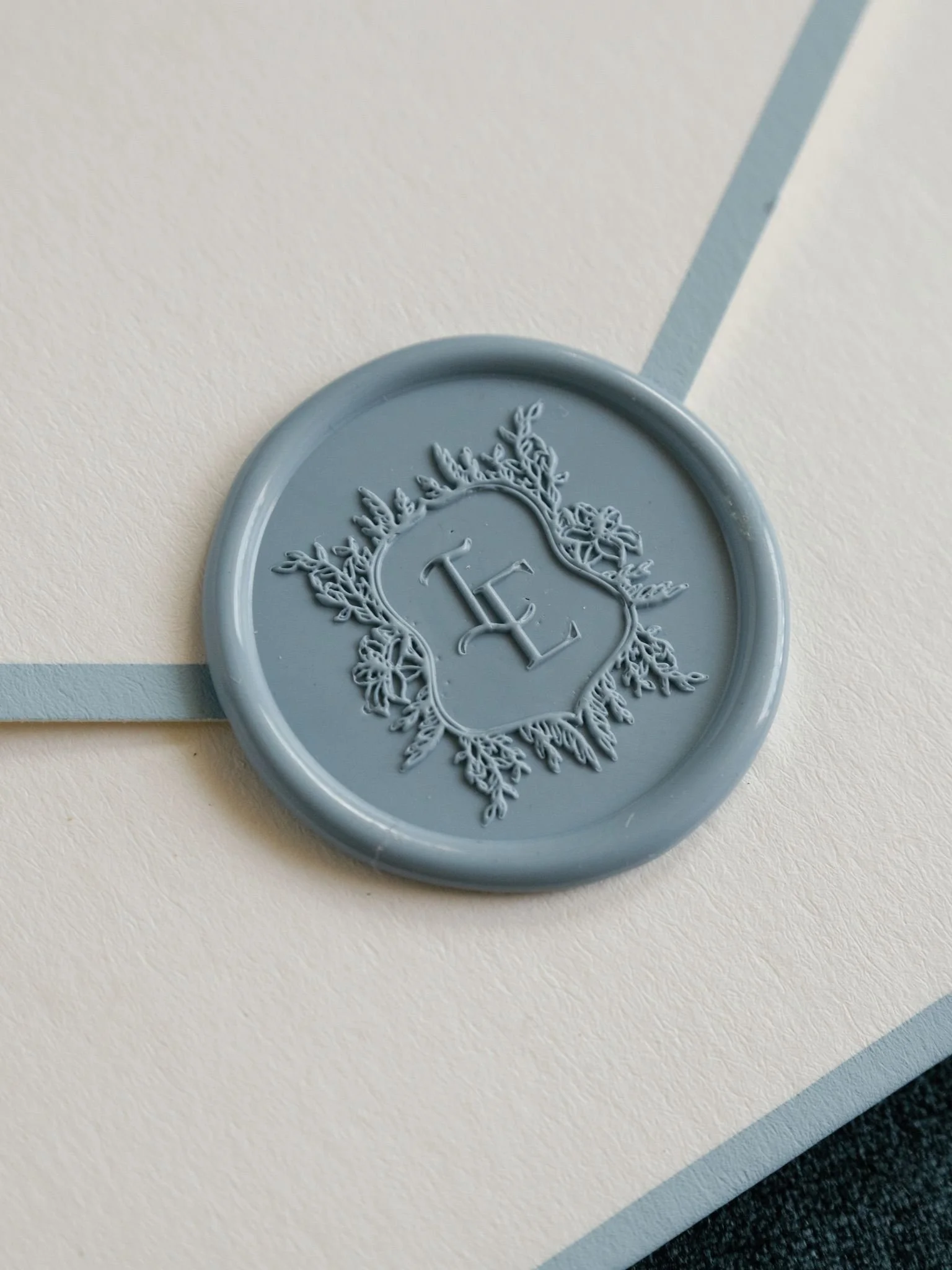 A round, light blue wax seal with an intricate floral design and the letter 'L' in the center, placed on a two-toned table.