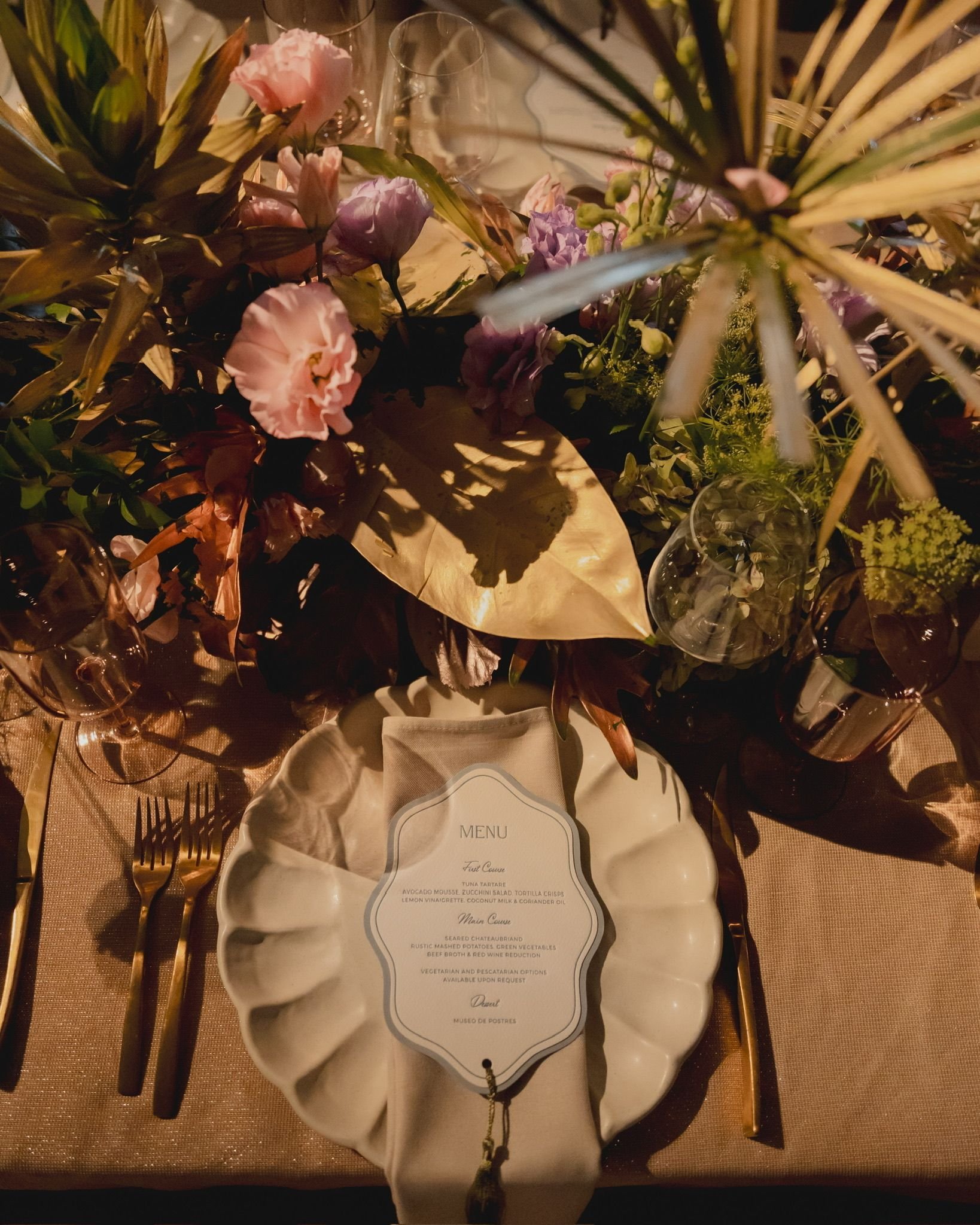 A table setting with golden utensils, a floral arrangement with pink flowers and greenery, and a menu card in the center. In the background, there are empty wine glasses and a glass of water.