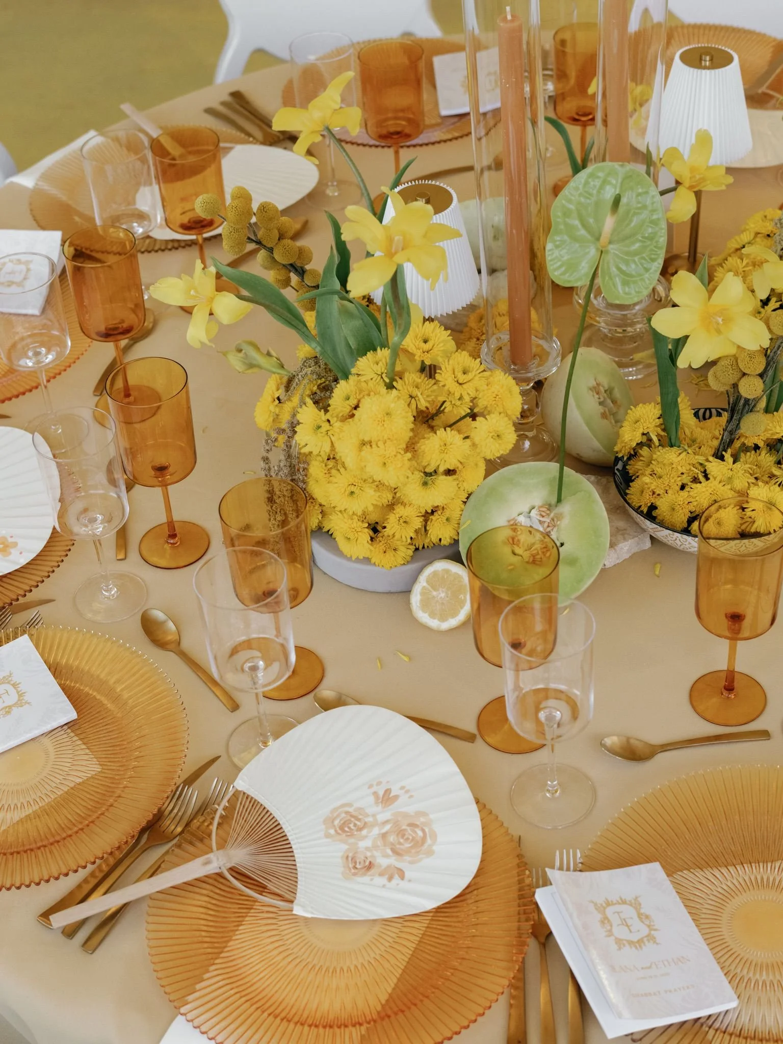 Table setting with yellow and orange color theme, featuring orange glasses, gold utensils, white and gold themed napkins, yellow floral centerpiece, and decorative paper fans.