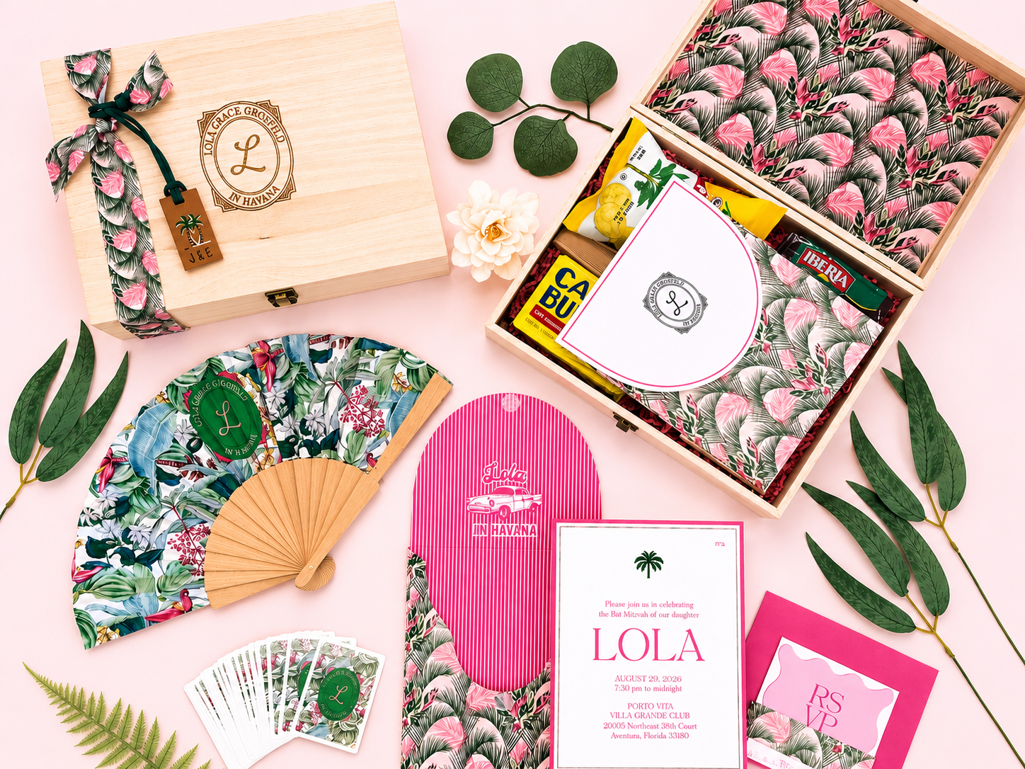 Tropical-themed party or gift set with wooden box, paper fans, floral stationery, green leaves, and small snack packs on a pink background.