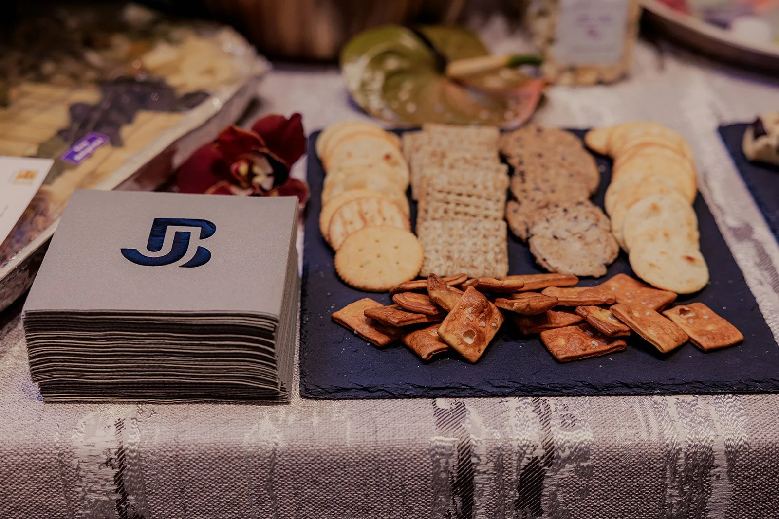 An assortment of crackers and pretzels on a black slate serving platter, with a stack of paper napkins with a stylized logo on the left, all on a textured tablecloth.