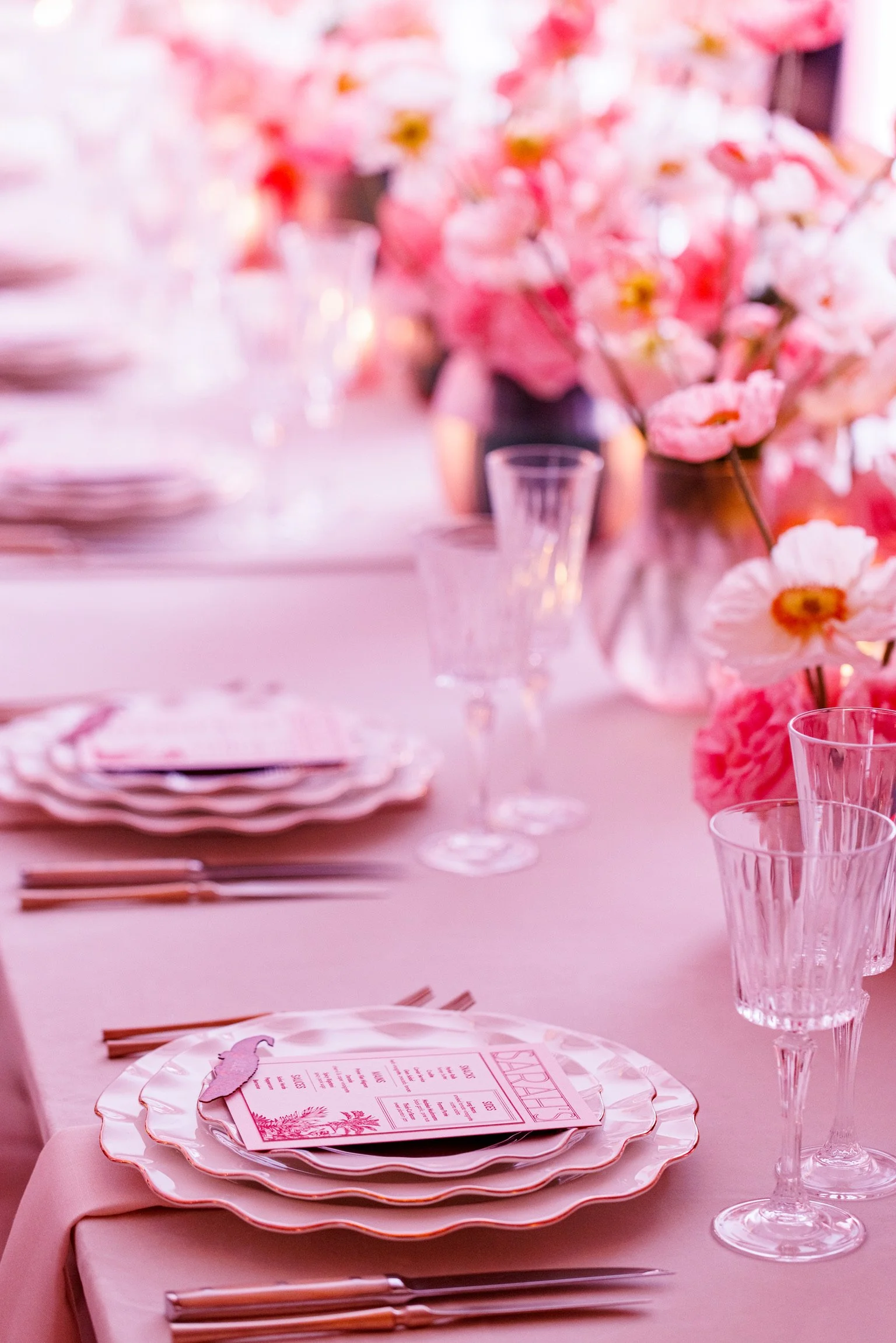 A pink-themed table setting with stacked plates,, menu cards, clear glasses, floral centerpieces with pink flowers, and pink napkins and utensils.