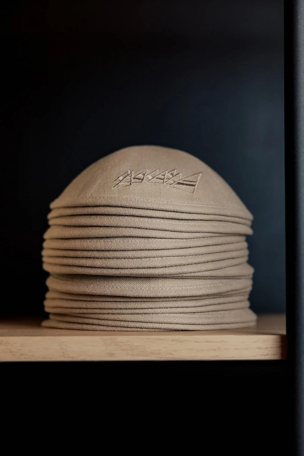Stack of beige cloth caps with embroidered logo on top, placed on a wooden shelf against a dark background.