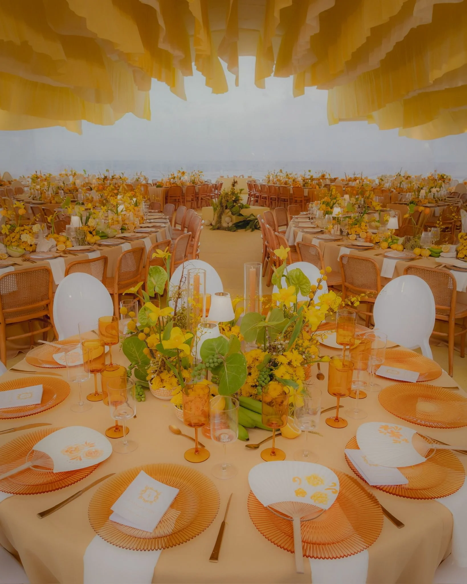 A daytime outdoor wedding reception setup on a beach with round tables arranged in a U-shape. The tables have light-colored tablecloths, white and orange paper plates, clear and orange glassware, and napkins with floral designs. The tables are decora