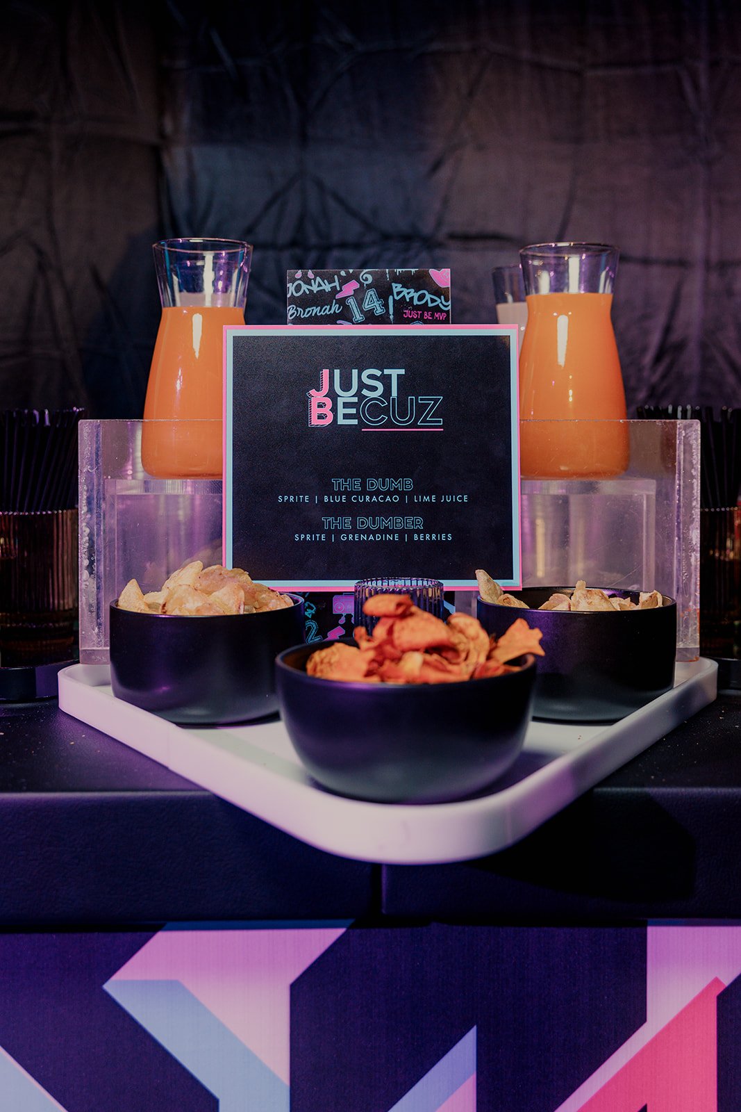 Buffet table with two orange juice pitchers, three bowls of snacks, and a menu with drink options labeled 'Just Because' at a party or event.