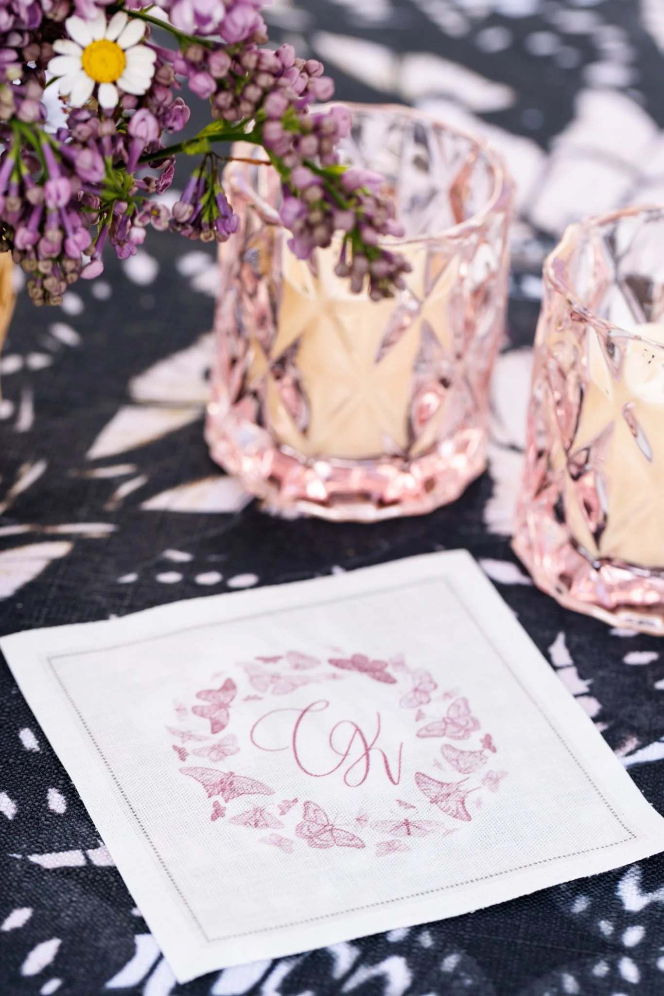 A table setting with a pink glass candle holder, a small bouquet of purple and white flowers in a yellow flower vase, and a paper napkin with a floral design and the word 'Love' written on it, all on a black and white patterned tablecloth.