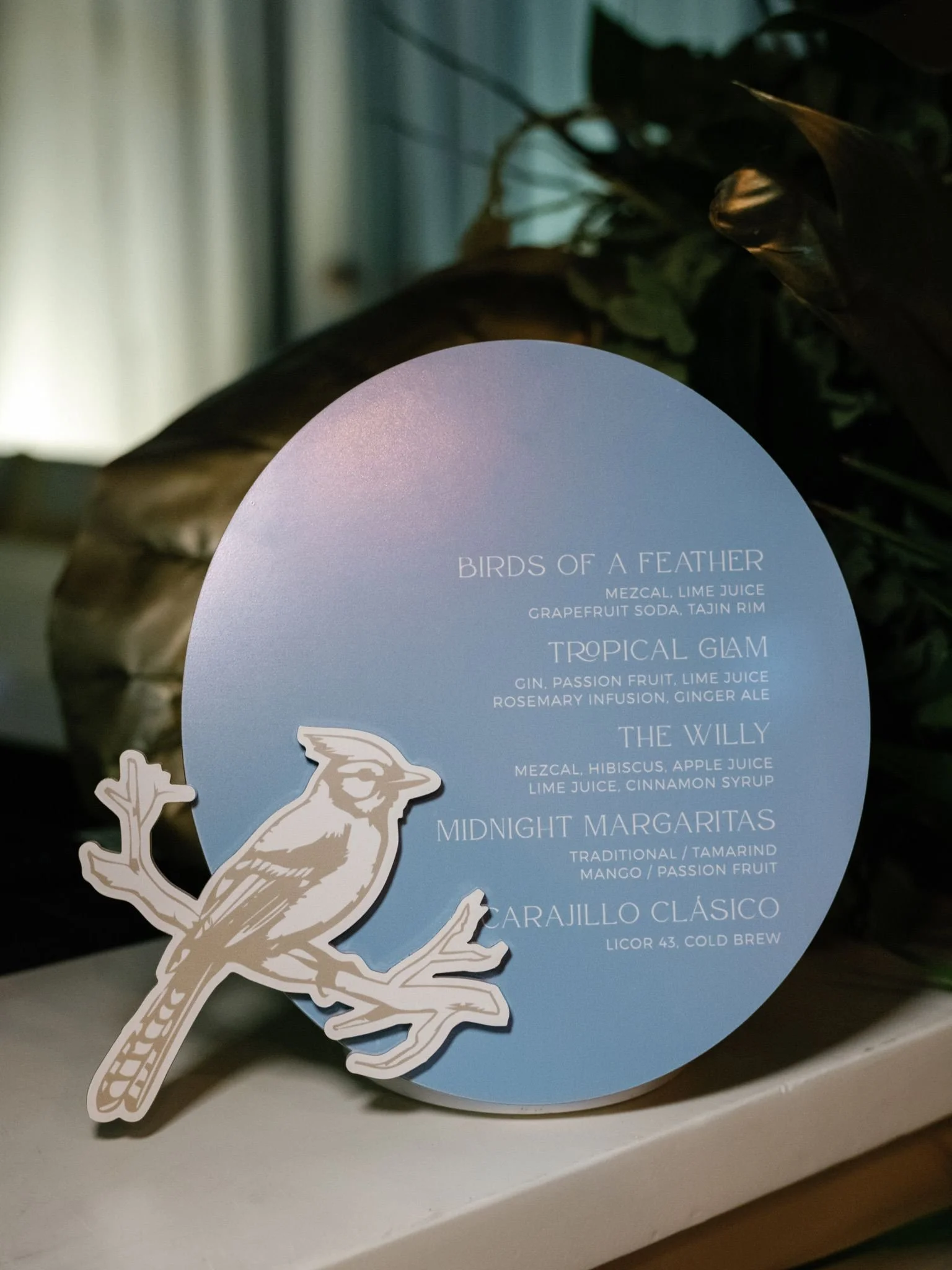 A circular drink menu with a bird illustration in the foreground and a list of cocktails and ingredients. The menu includes drinks such as Birds of a Feather, Tropical Glam, The Willy, and Midnight Margaritas, with ingredients like mezcal, lime juice