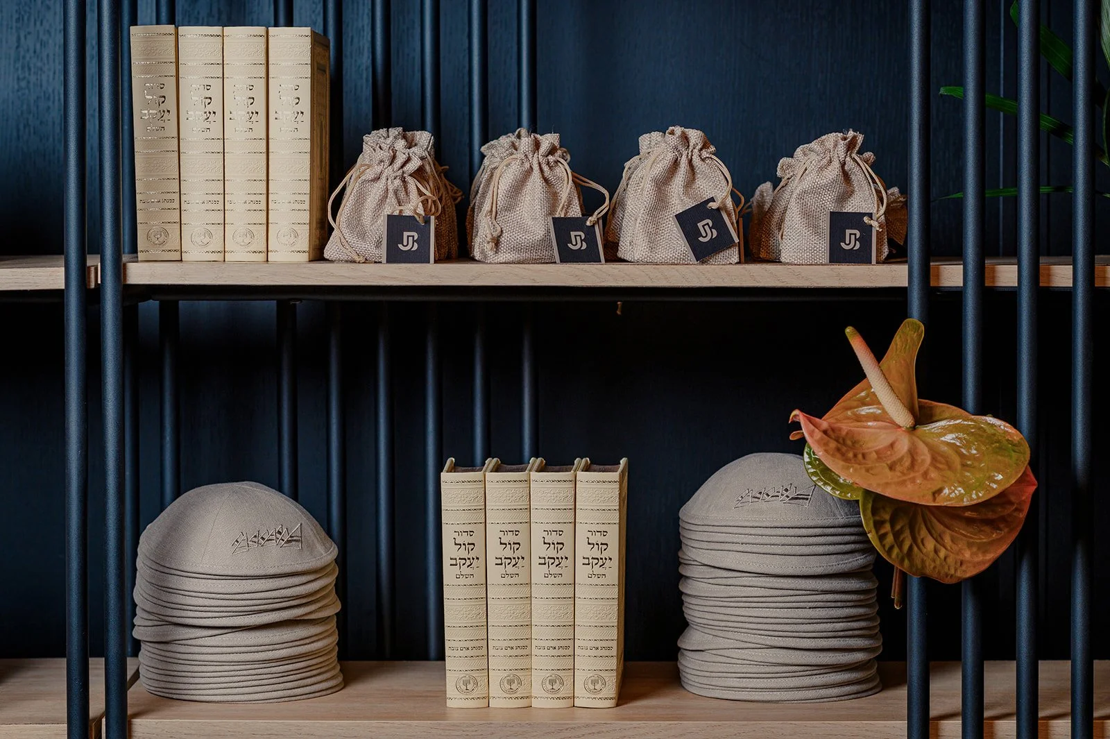 Shelf with beige books, small cloth pouches with tags, beige head coverings, and an anthurium flower.