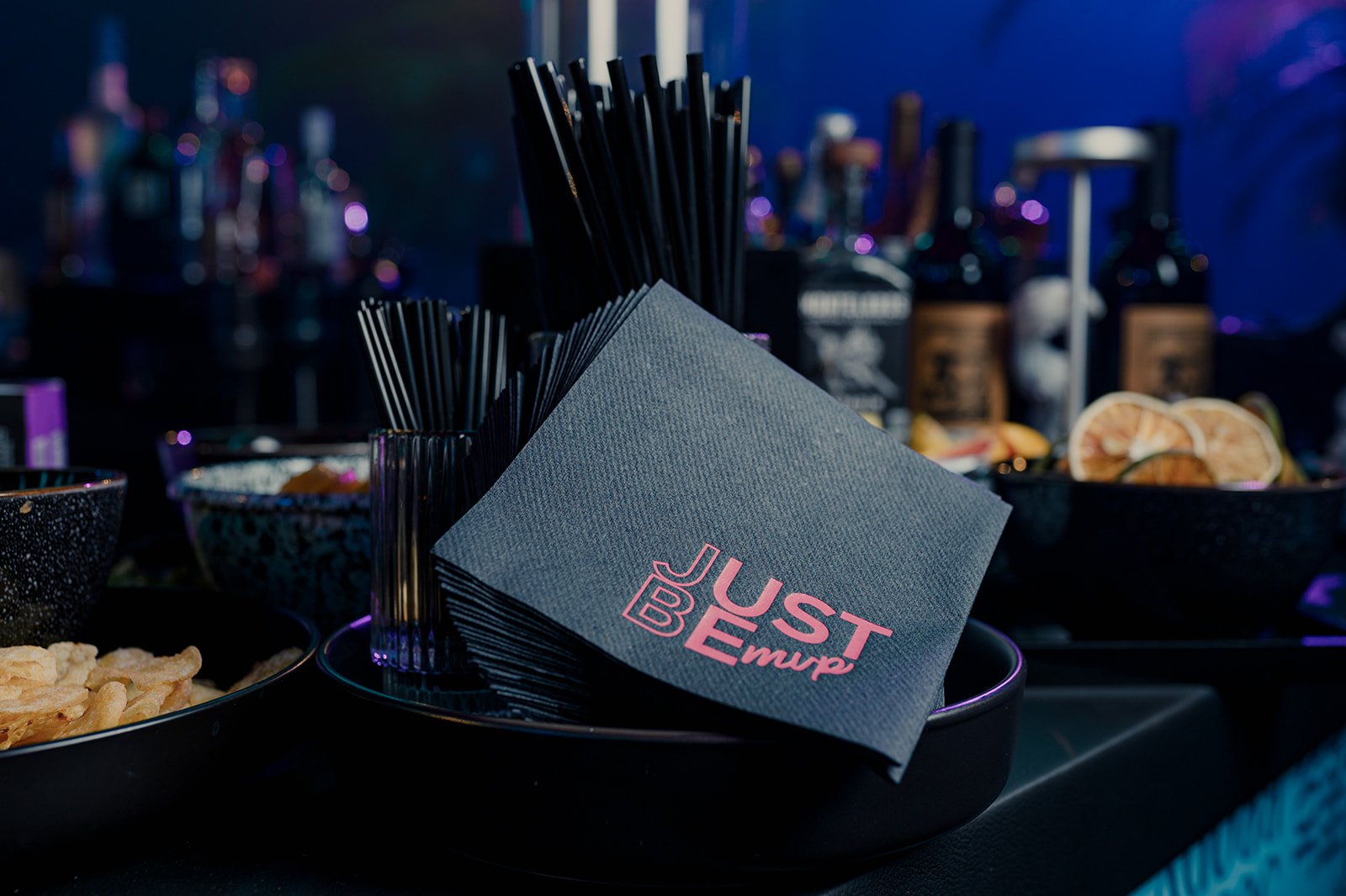 Black napkin with pink text that reads 'Just Be Emp' next to black straws on a black tray, with bowls of snacks and a bar in the background.