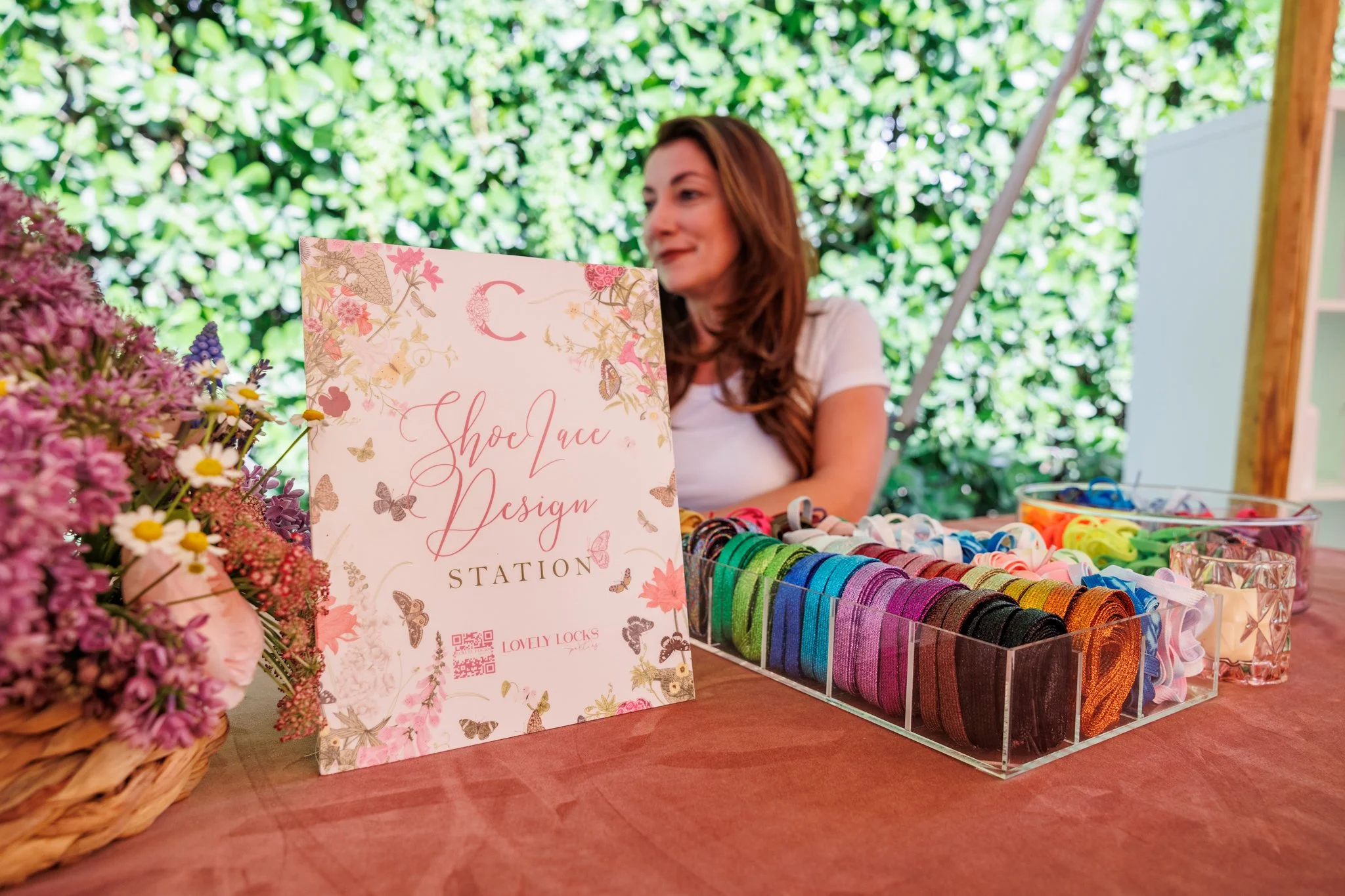 Woman sitting behind a display of colorful ribbons at a craft or accessory booth, with a floral sign that reads 'Shoe Lace Design Station' in front and a basket of pink flowers to the side, outside under a shaded, leafy canopy.