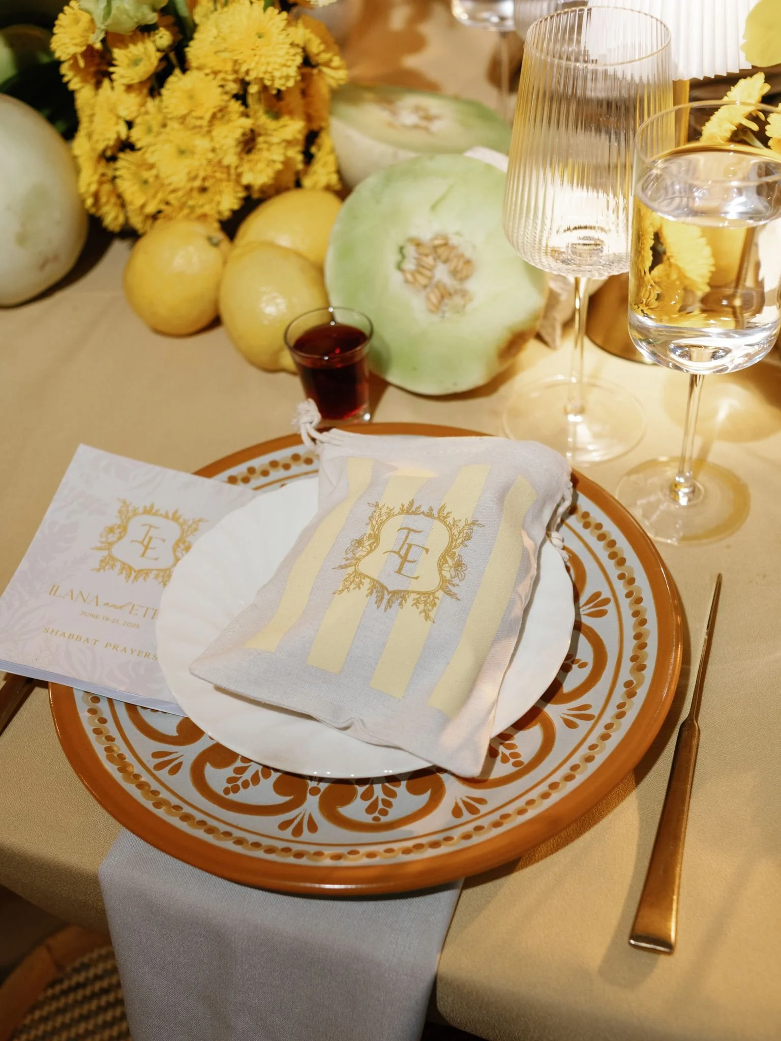 A table setting with a decorative plate, paper napkins with gold accents, yellow flowers, a sliced honeydew melon, three yellow apples, a small glass of red juice, and two glasses of water. The table is covered with a beige tablecloth.