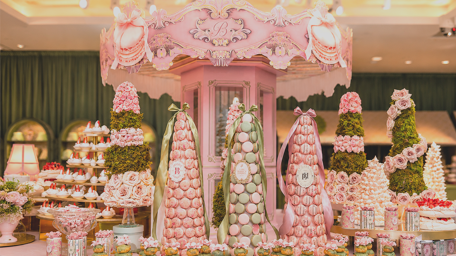 Pink and green floral-themed dessert display with macaron towers, decorated with ribbons, roses, and other confections, under a pink decorative canopy.