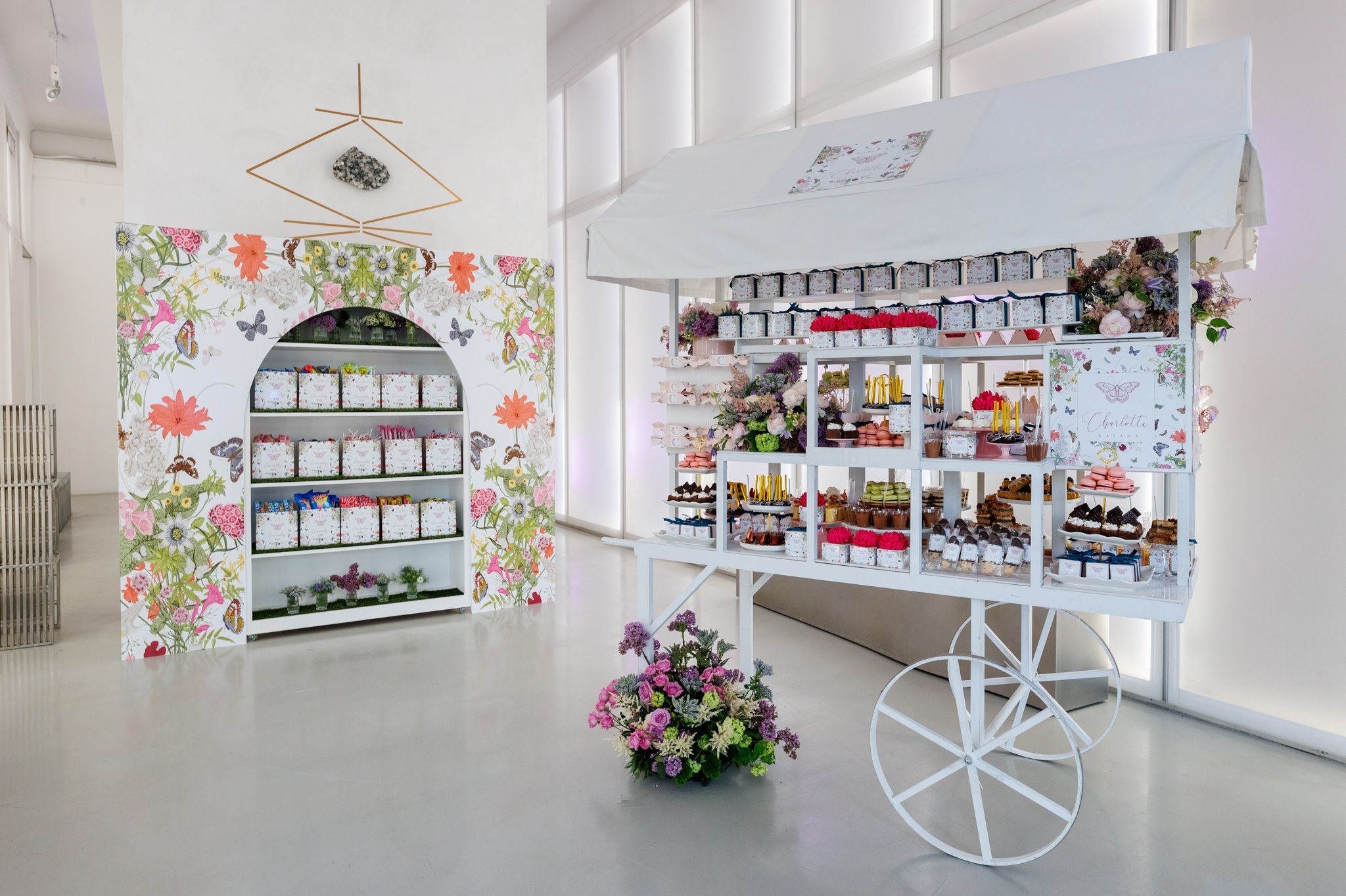 Photo of a white dessert cart with flowers on it, and a white floral backdrop with a floral arch and shelves of favors and sweets inside a bright room with large windows.