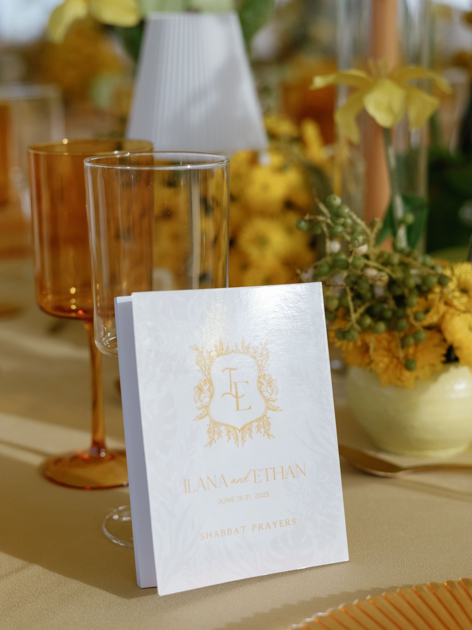 A wedding program card standing on a table, with wine glasses and floral arrangements blurred in the background.