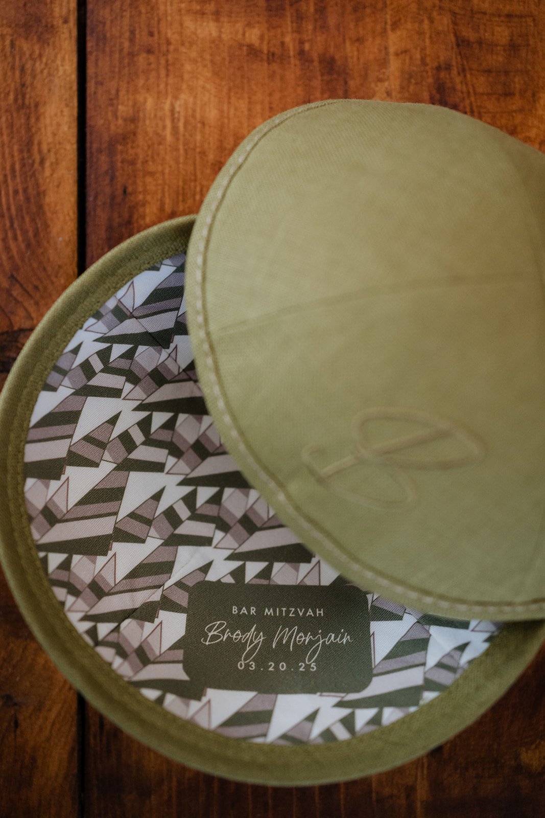 Open circular box with a green lid and patterned interior, containing a printed invitation for a Bar Mitzvah celebration for Brody Morgan on March 20, 2025.