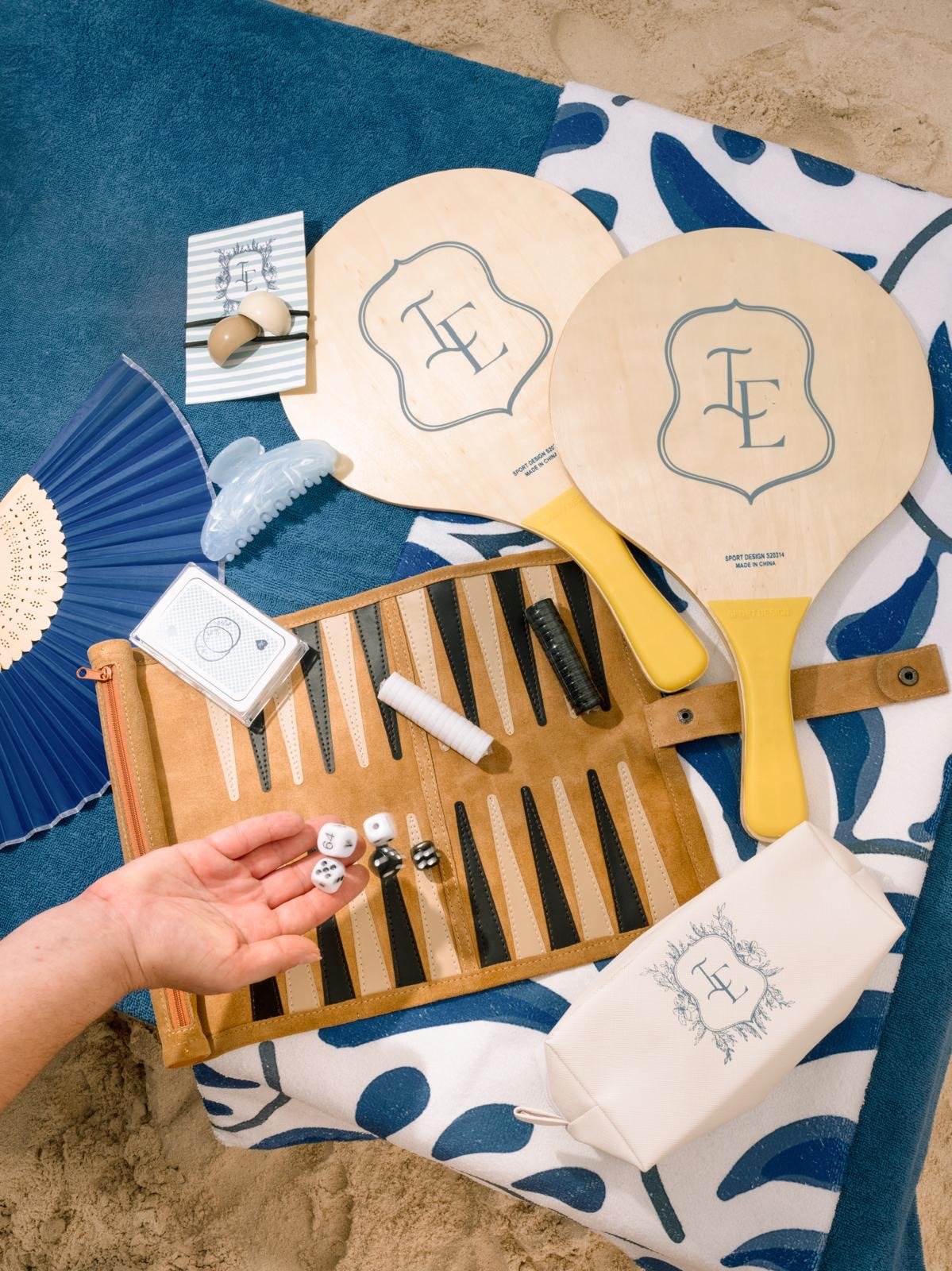 Beach scene with two yellow paddles, a brown roll-up bag containing black and white dominoes, dice, and small game pieces, a blue and white patterned towel, a blue fan, decorative black and white hair clips, and other beach accessories.
