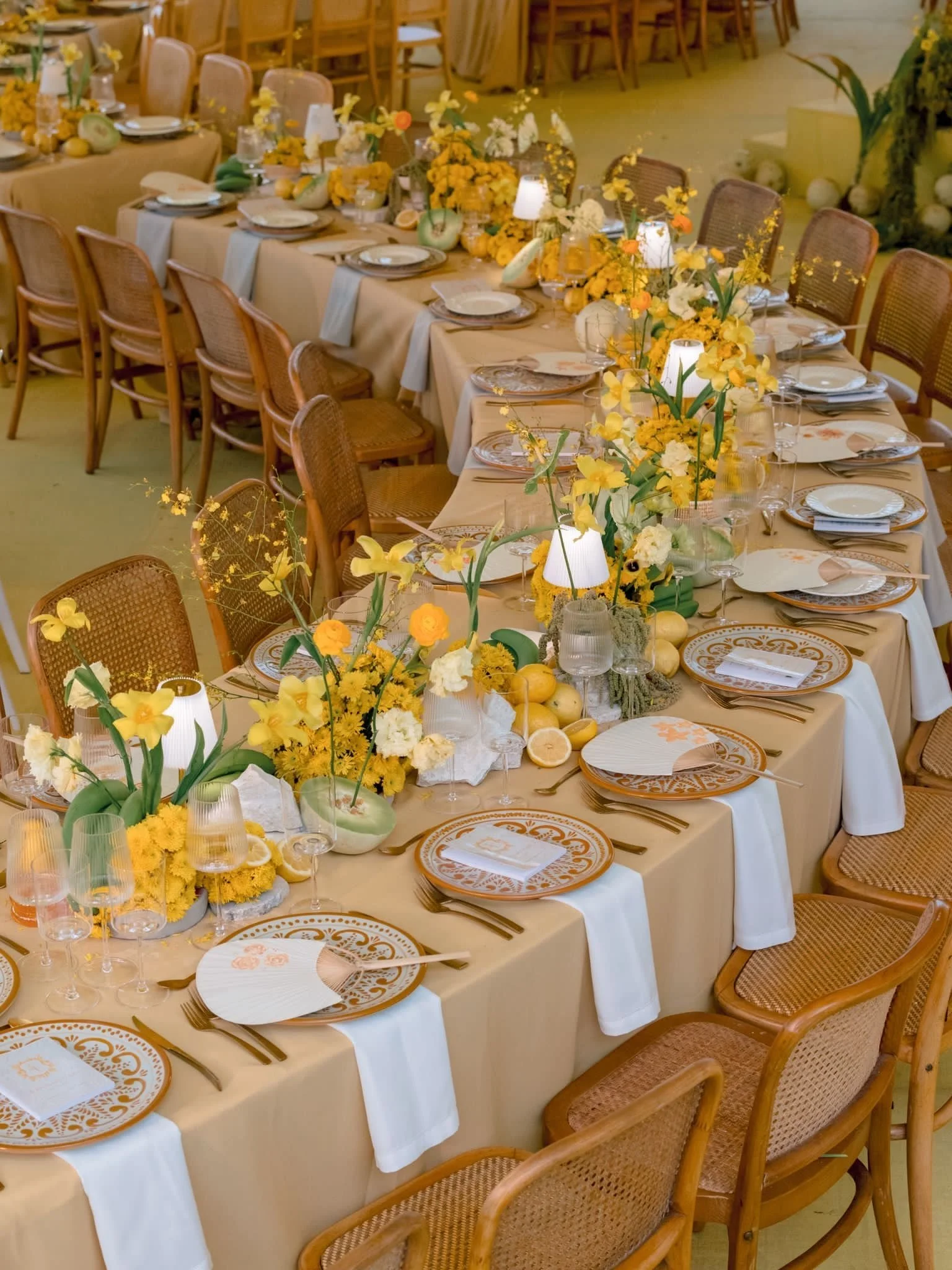A long banquet table decorated for a celebration with yellow and white flowers, yellow candles, lemons, and place settings with white napkins and ornate orange and white plates. There are wicker chairs with wood frames surrounding the table.
