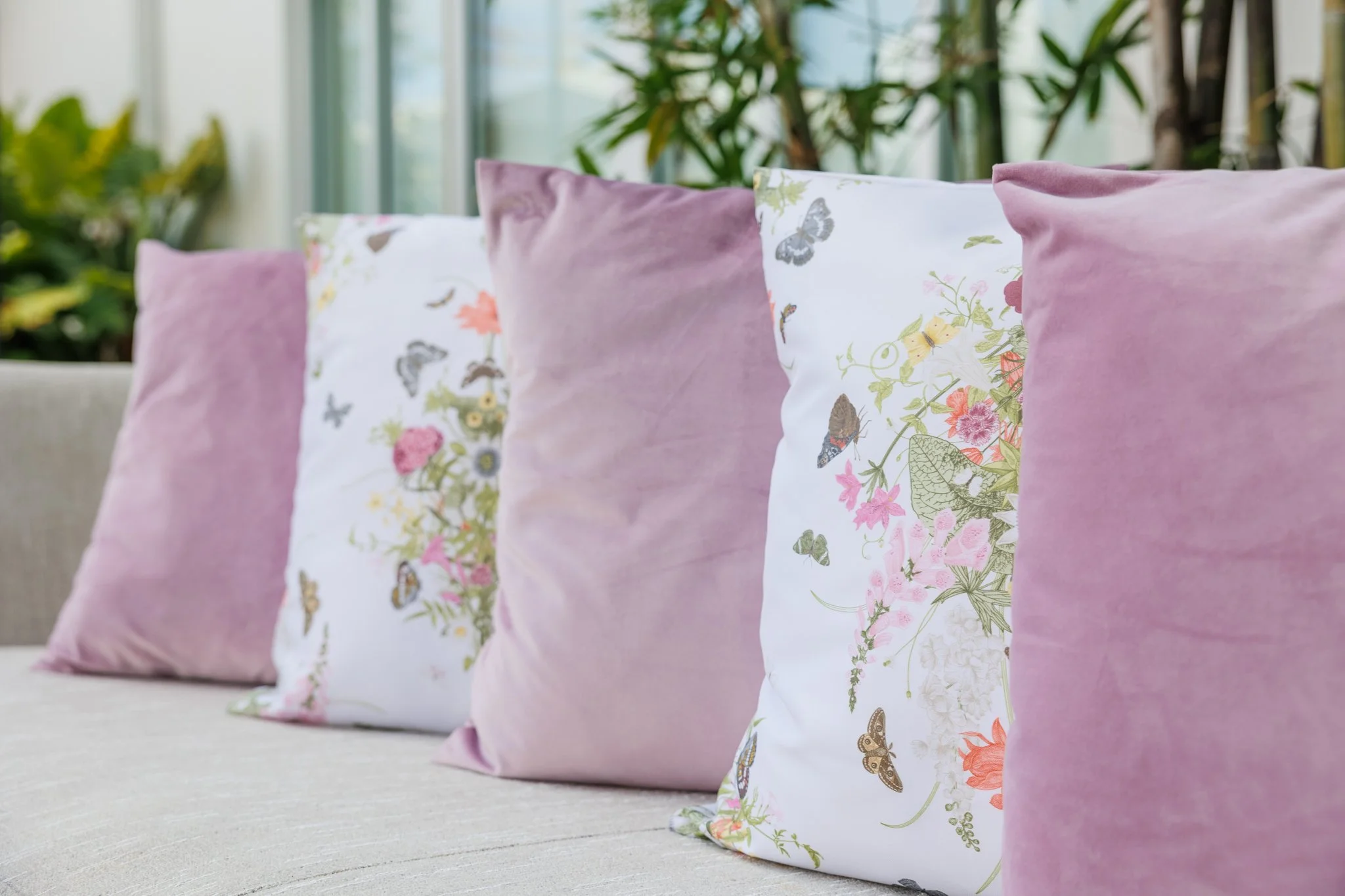 A row of decorative pillows on a sofa, with some pillows in pink velvet and others with colorful butterfly and floral print covers, set in a bright room with large windows and greenery outside.