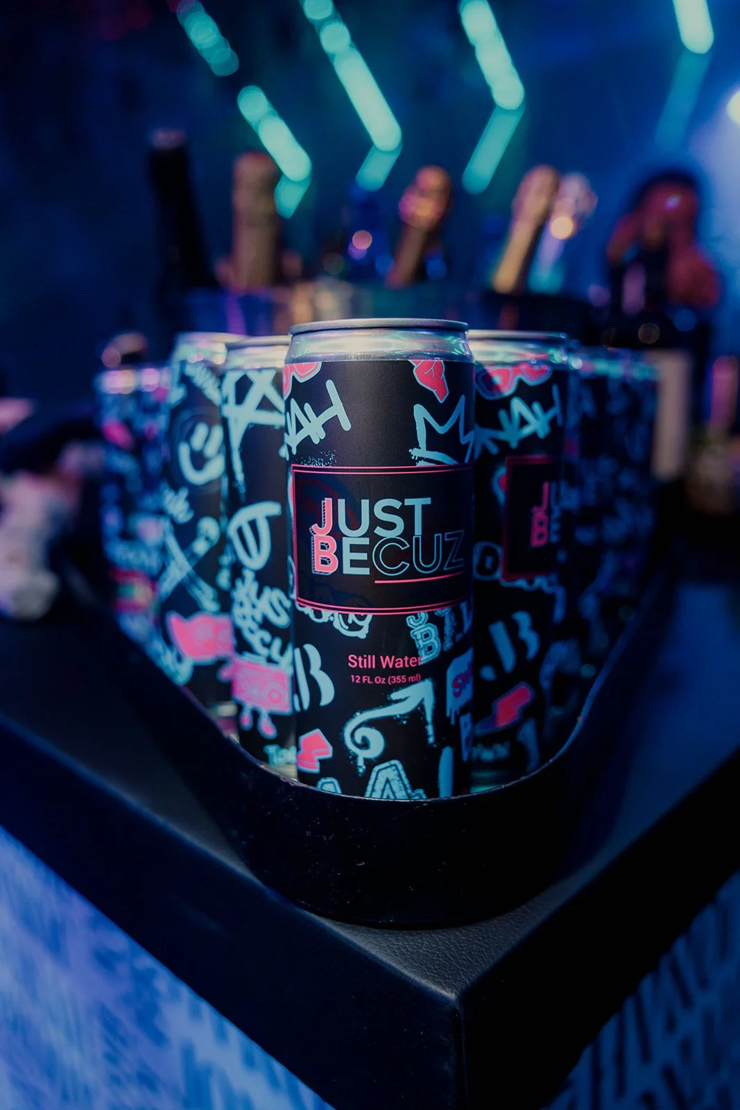 A can of water with graffiti-style design and the words 'Just Because' in a neon-lit setting, with multiple cans in the background.