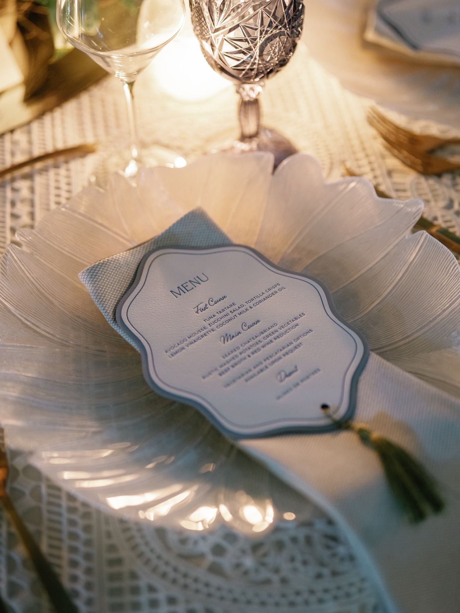 Elegant table setting with a menu, crystal glasses, and a white decorative plate, illuminated by a candle, on a lace tablecloth.