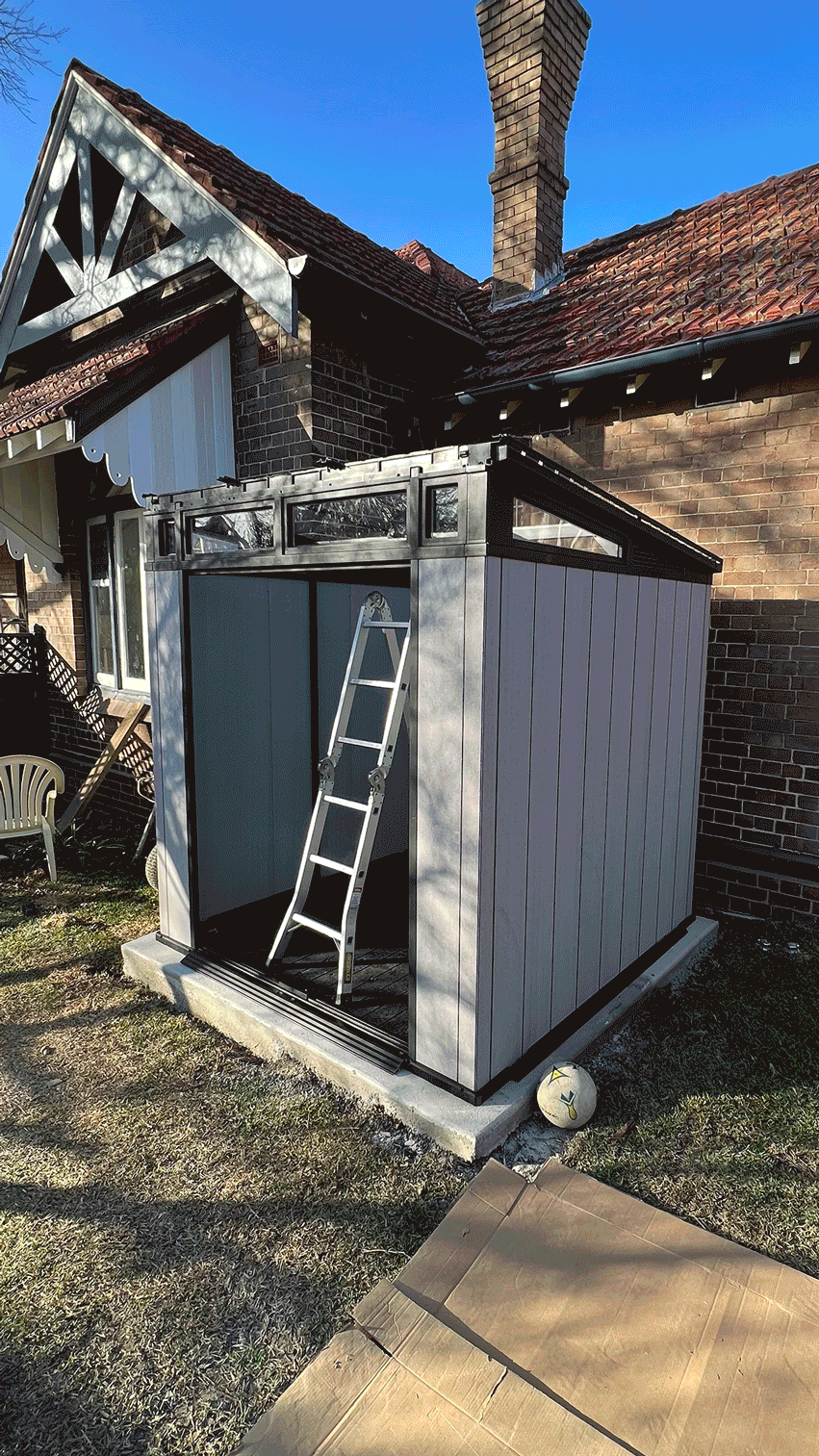 A small outdoor shed with a ladder leaning against it, situated on a concrete foundation outside a brick house. The shed has vertical panel siding and a slanted roof with windows at the top. Nearby there is a soccer ball and some cardboard on the ground.