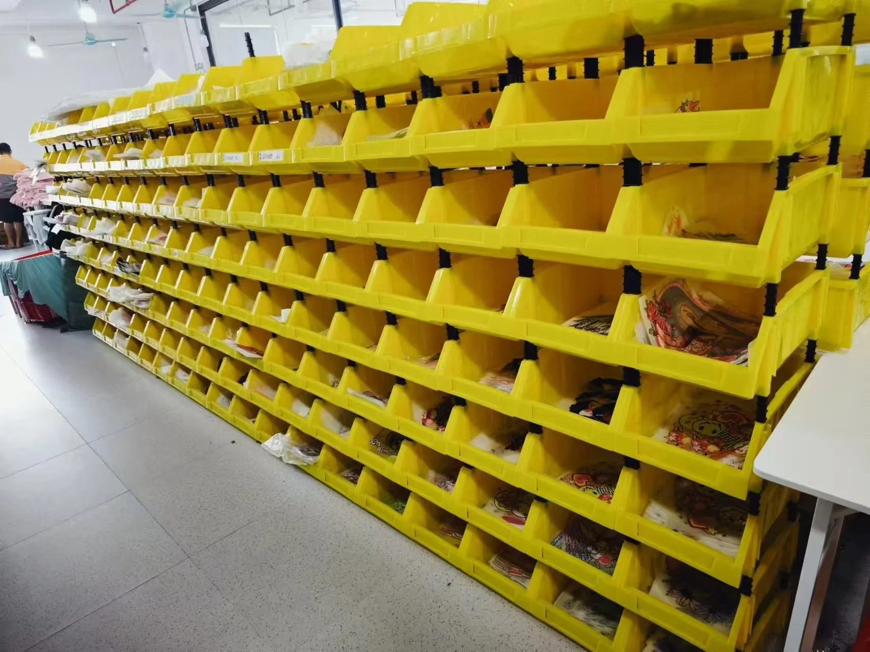 Yellow plastic storage bins with small black clips holding them together, filled with colorful graphics for fabric transfers on a metal shelving unit in an indoor setting.
