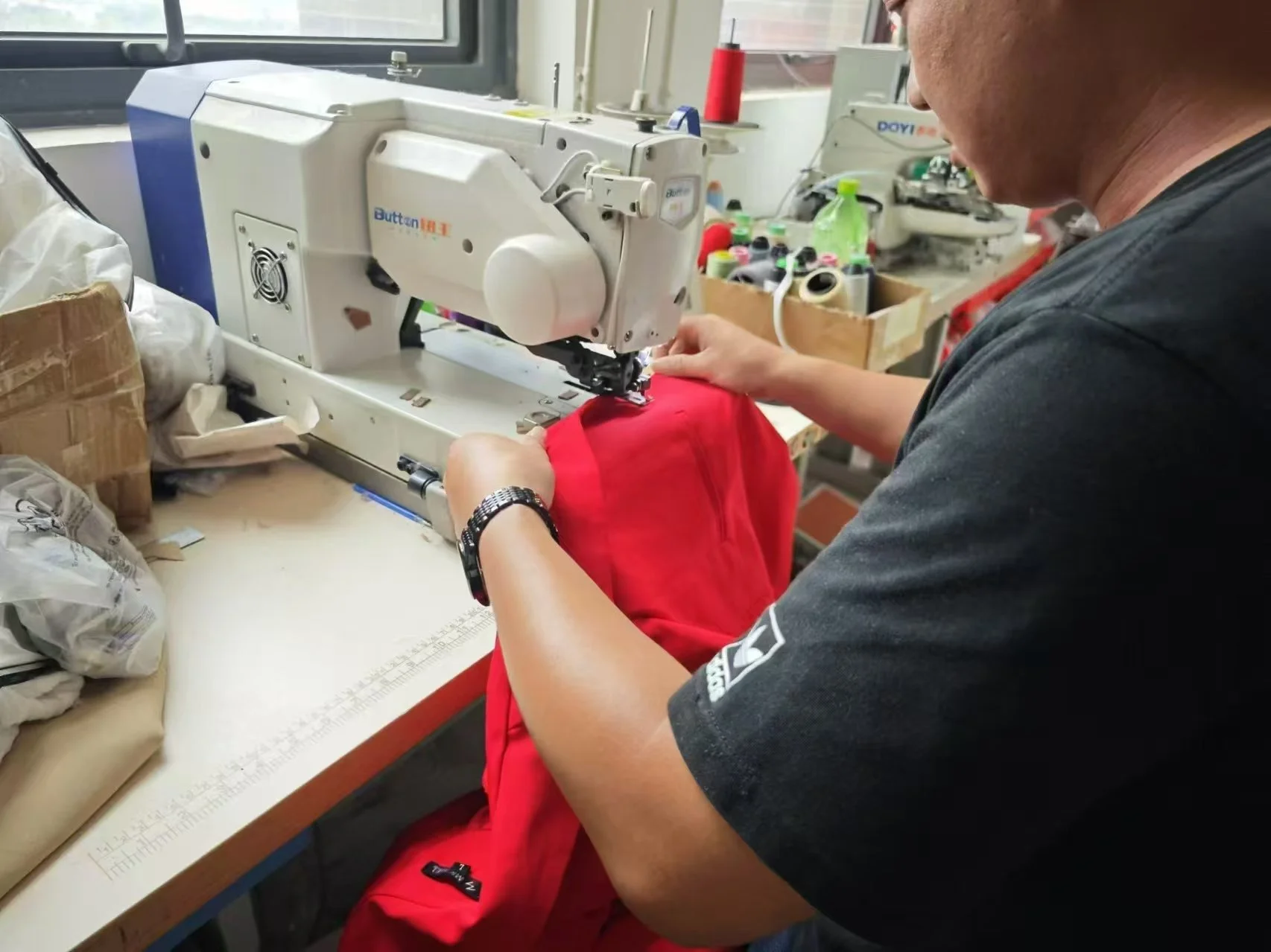 A person sewing a red fabric at a worktable in a workshop, with sewing machine and supplies visible.
