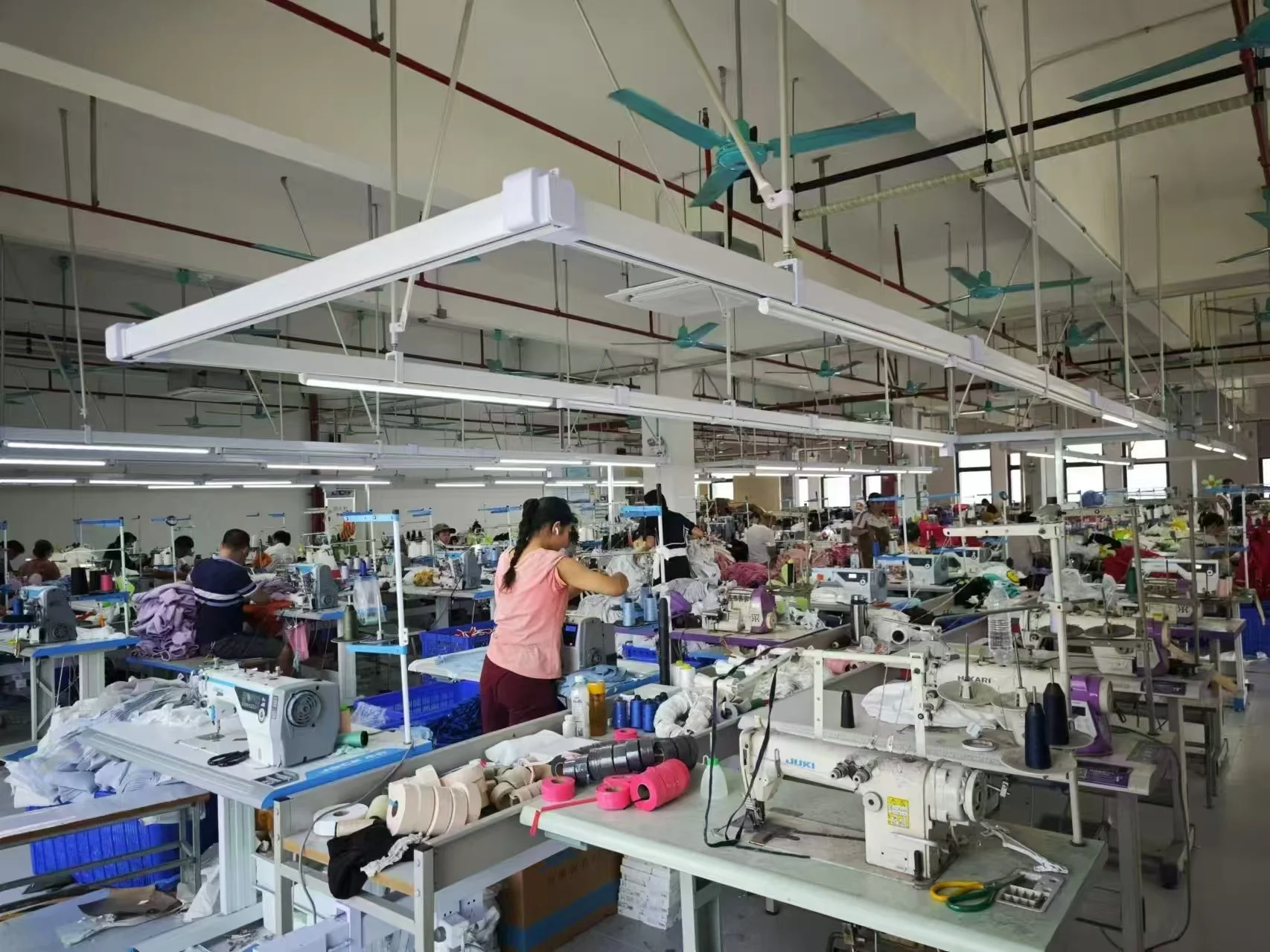A large factory or workshop with many workers at sewing machines, handling fabrics and sewing garments. The space is well-lit with fluorescent lights and ceiling fans, with industrial equipment on the tables and a spacious layout.