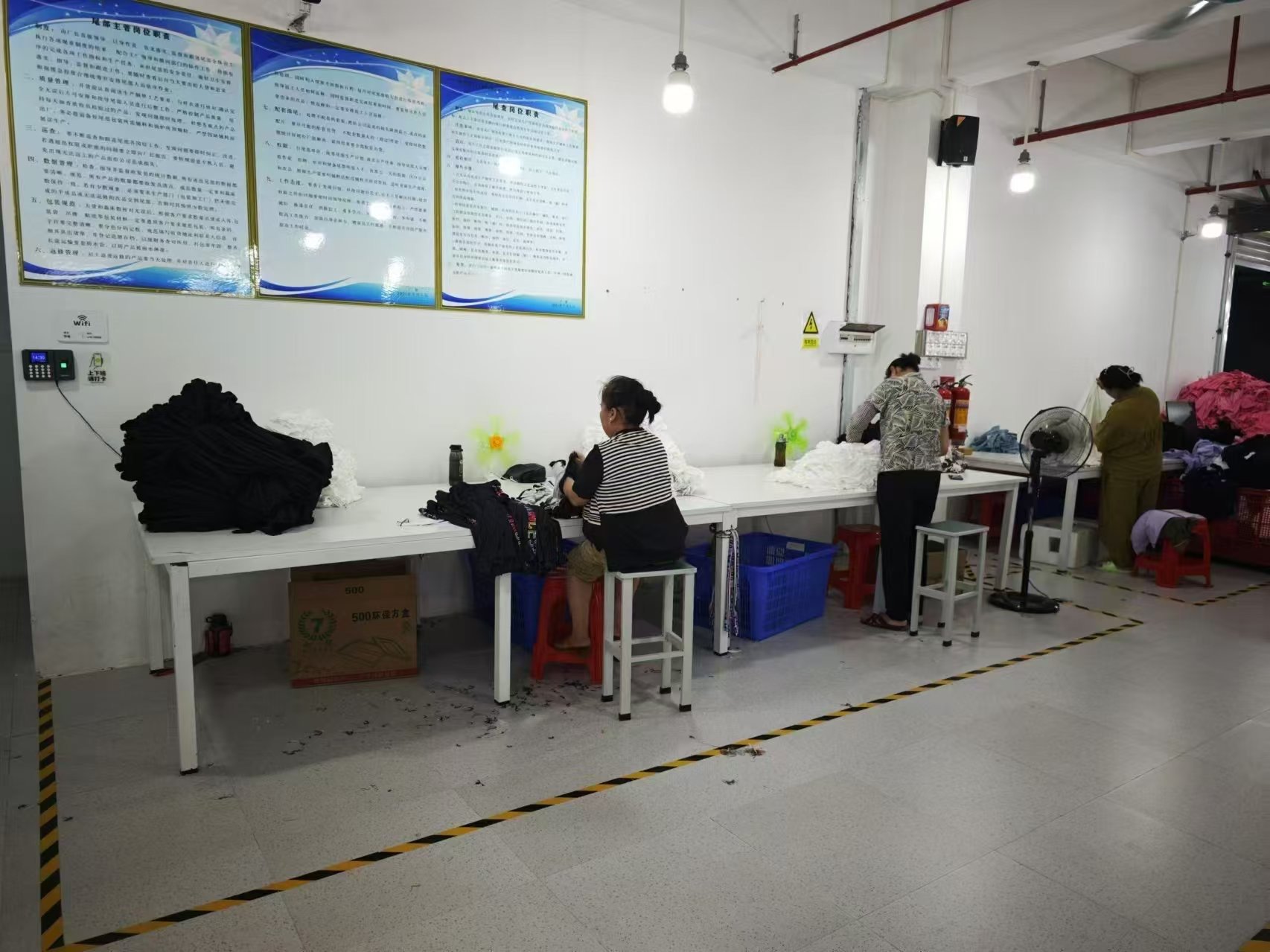 People working at a long white table with piles of fabric, in what appears to be a textile or clothing workshop; three women are working, with tools and bottles beside them, and the wall has three large informational posters with Chinese text.