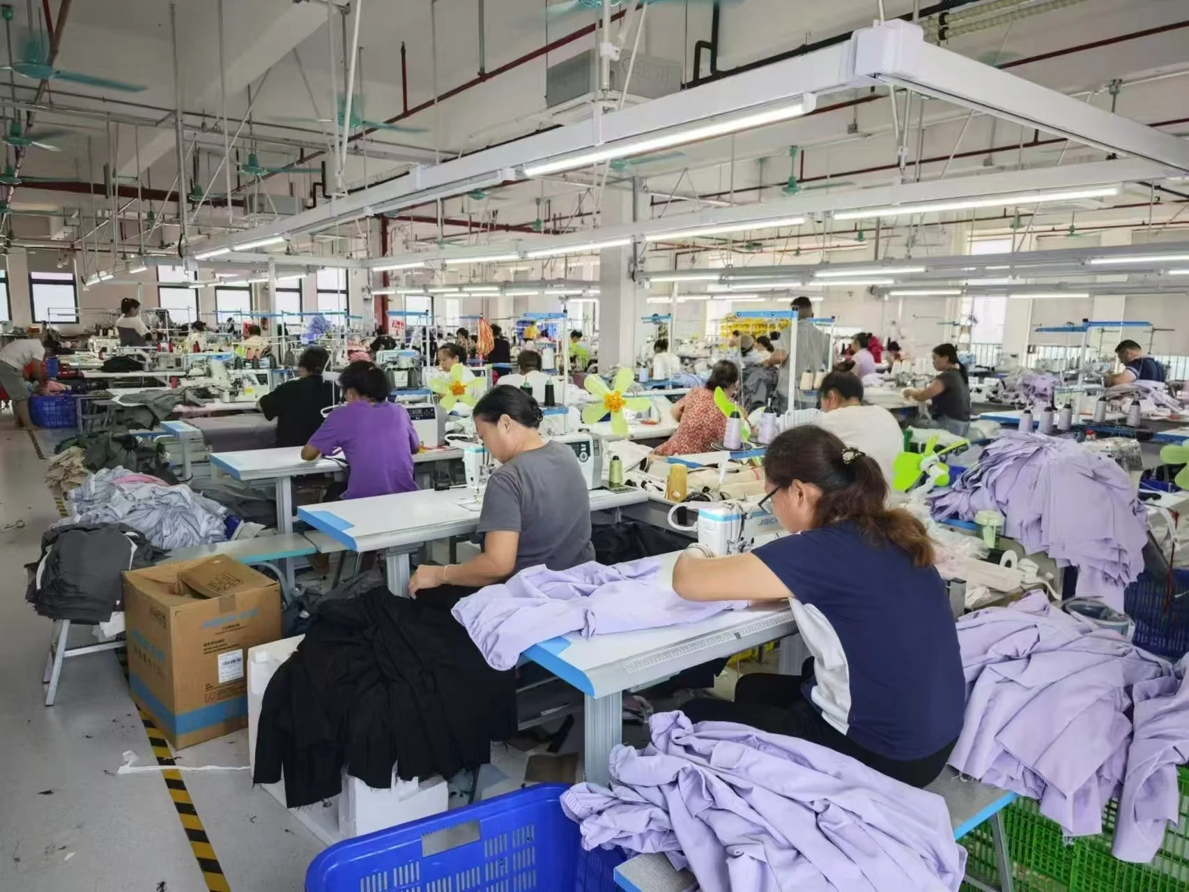 A busy garment factory with workers sewing clothes at long workstations. Piles of fabric and garments are on the tables, with some workers focusing on their tasks. The factory has high ceilings, fluorescent lighting, and large windows, with more work