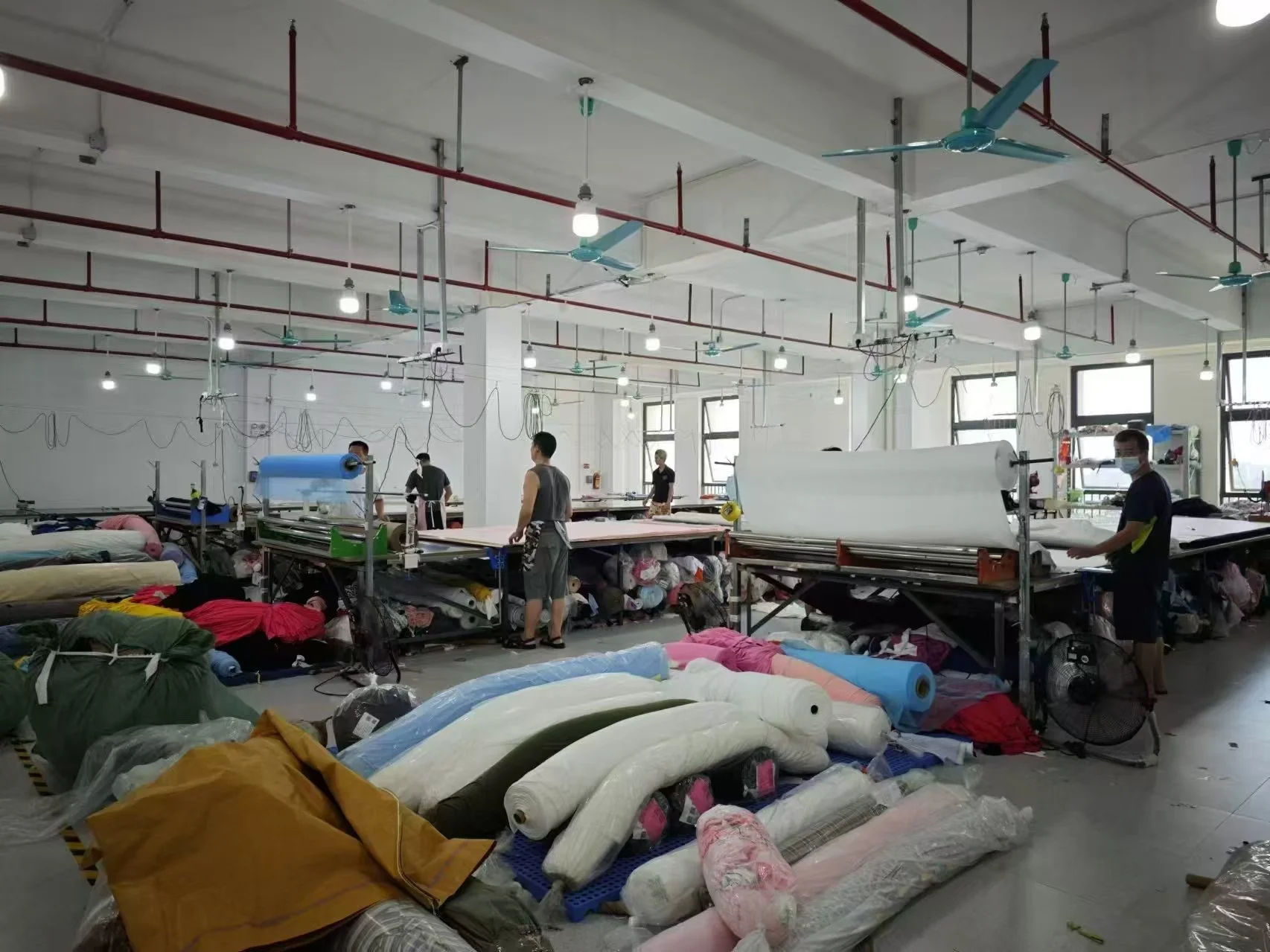 Workers in a textile or fabric factory working at cutting tables amid rolls of fabric and fabric scraps, with sewing machinery, fans, and ceiling fans overhead.