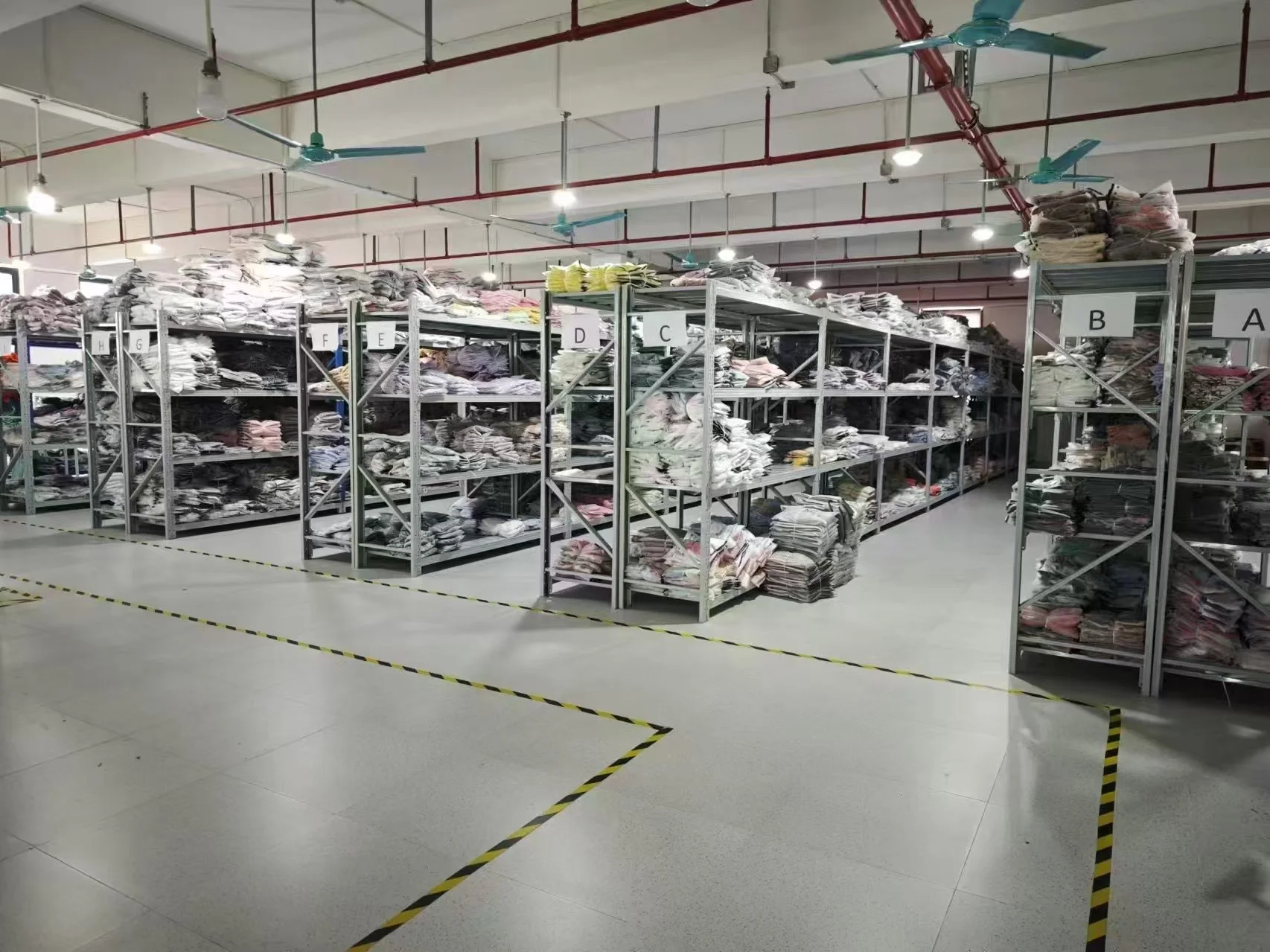 A warehouse with metal shelves stacked with folded clothes, labeled with alphabetic tags, under bright ceiling lights and ceiling fans, with marked floor sections.