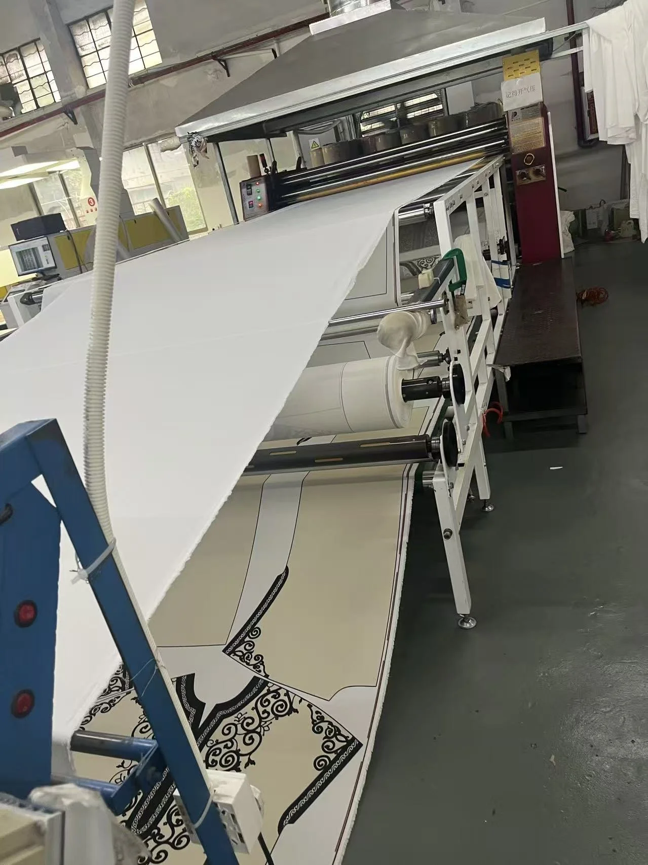 Industrial printing machine printing decorative black and white tile patterns on large sheets of paper or fabric inside a factory.