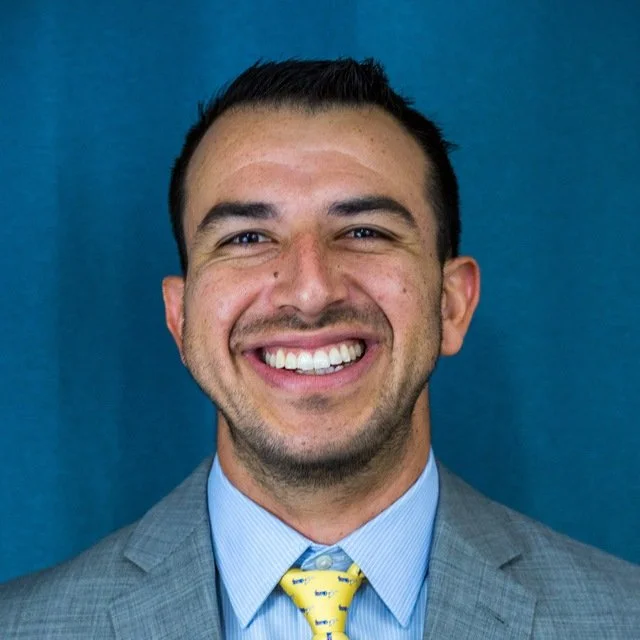 Professional man with short dark hair, smiling, wearing a gray suit, light blue shirt, and yellow tie with blue patterns, standing against a blue background.