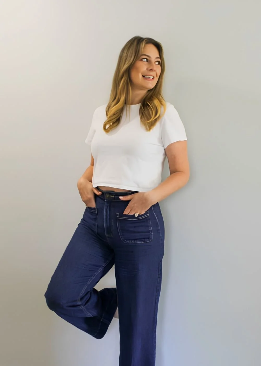 Maddy Cosgrove, wavy blonde hair wearing a white t-shirt and dark blue jeans, standing against a plain light gray wall, smiling and looking to her right with one hand in her pocket.