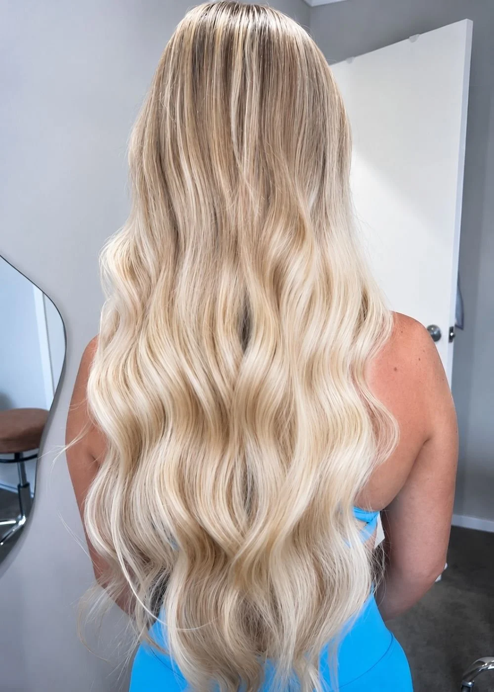 Back of a woman with long, wavy blonde hair in a salon or studio with a mirror and white door in the background.