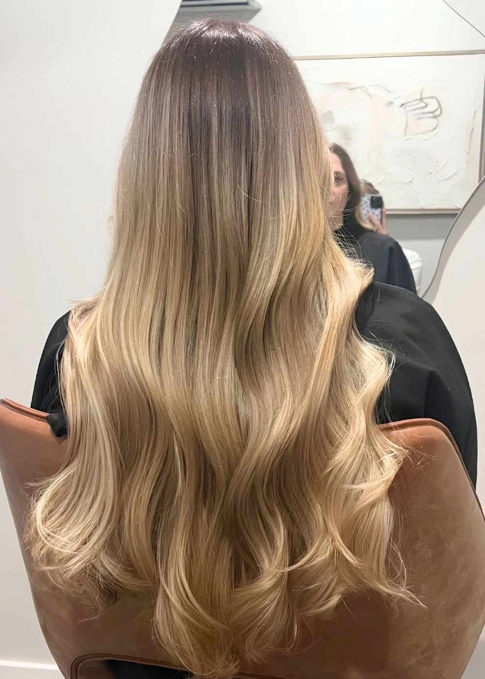 Back view of a woman with long, wavy blonde hair sitting in a salon chair, reflected in a mirror.