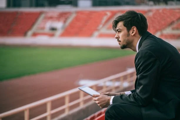 A man in a suit sitting on a bench at a sports stadium, looking at a tablet. The stadiums seats are visible in the background.