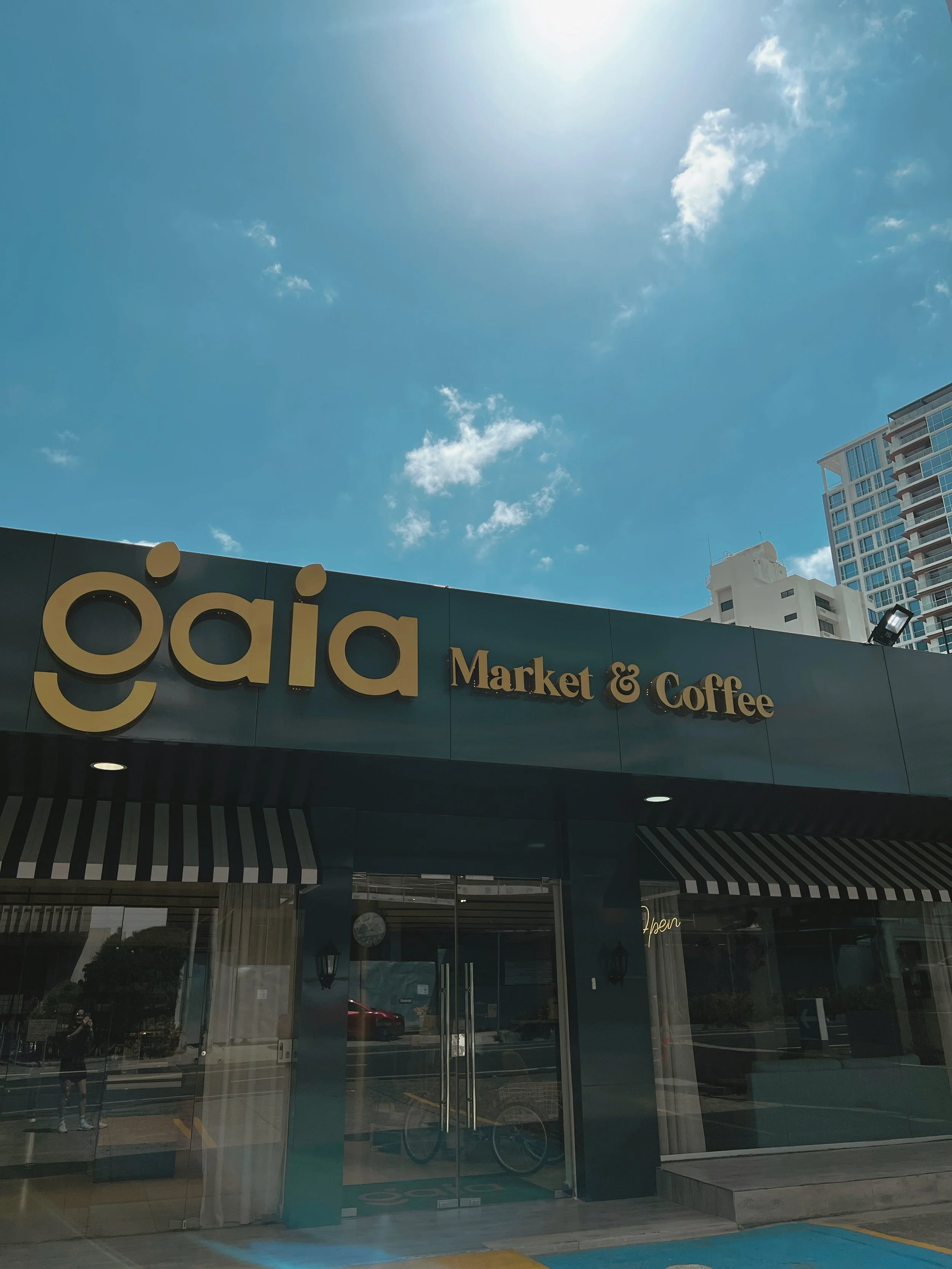 Entrada de una tienda llamada 'gāia Market & Coffee' con letras en color dorado sobre un fondo azul, con puertas de vidrio y una tienda en el interior visible a través del vidrio, y edificios altos y cielo azul con algunas nubes en el fondo.