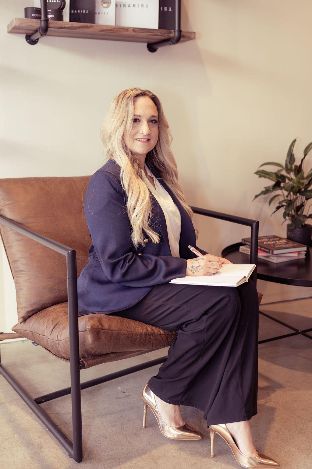A woman with long wavy blonde hair, wearing a navy blazer, white blouse, black pants, and gold high heels, sits on a brown leather chair in an office setting. She is smiling, holding a pen, and has a notebook on her lap, with a plant and books on a side table behind her.
