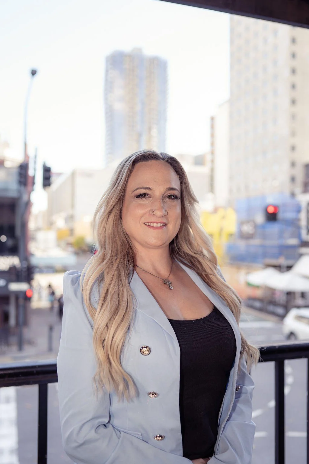 A woman with long, wavy blonde hair standing outdoors in an urban setting with tall buildings and busy street behind her.