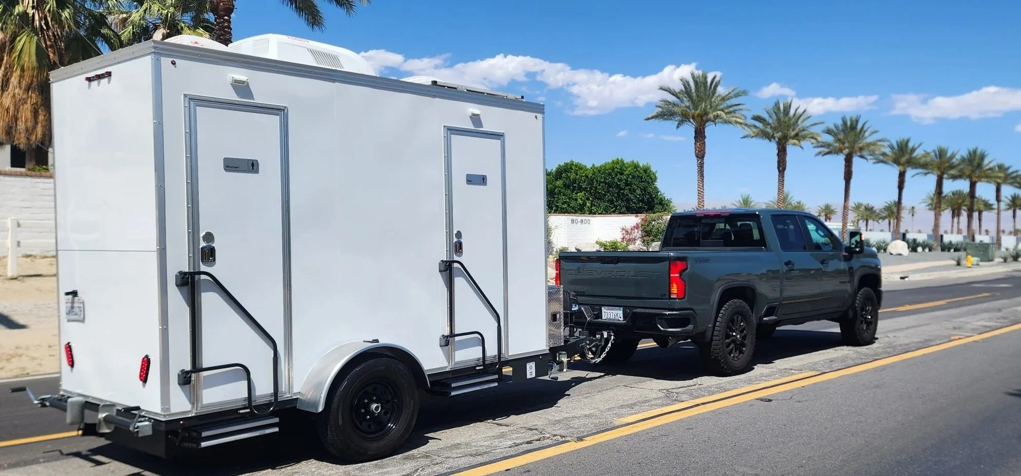 A black Chevrolet pickup truck towing a white mobile restroom trailer parked on a sunny street lined with palm trees.