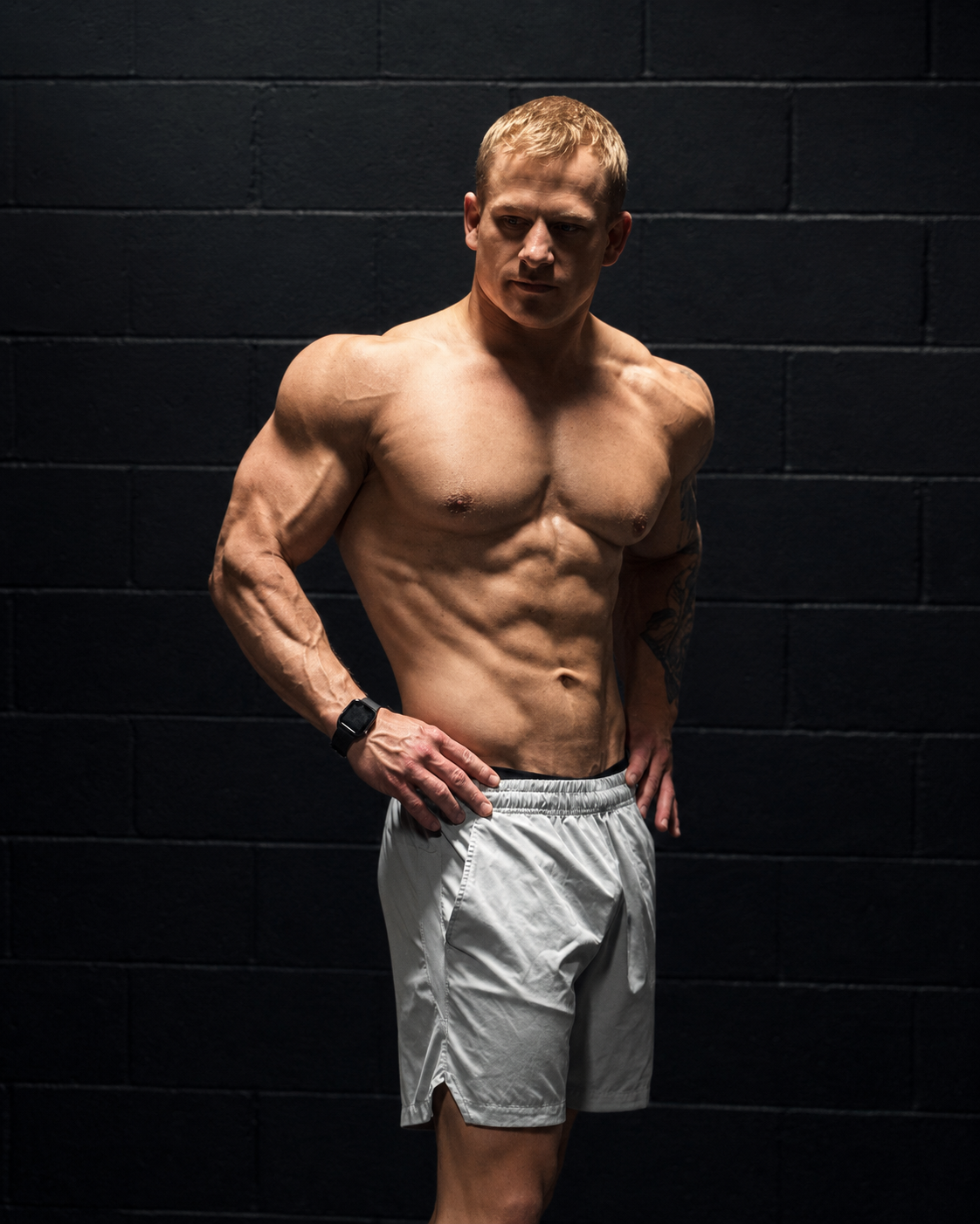 A shirtless man with muscular build and tattoos stands against a black brick wall, looking to the side with his hands on his hips.