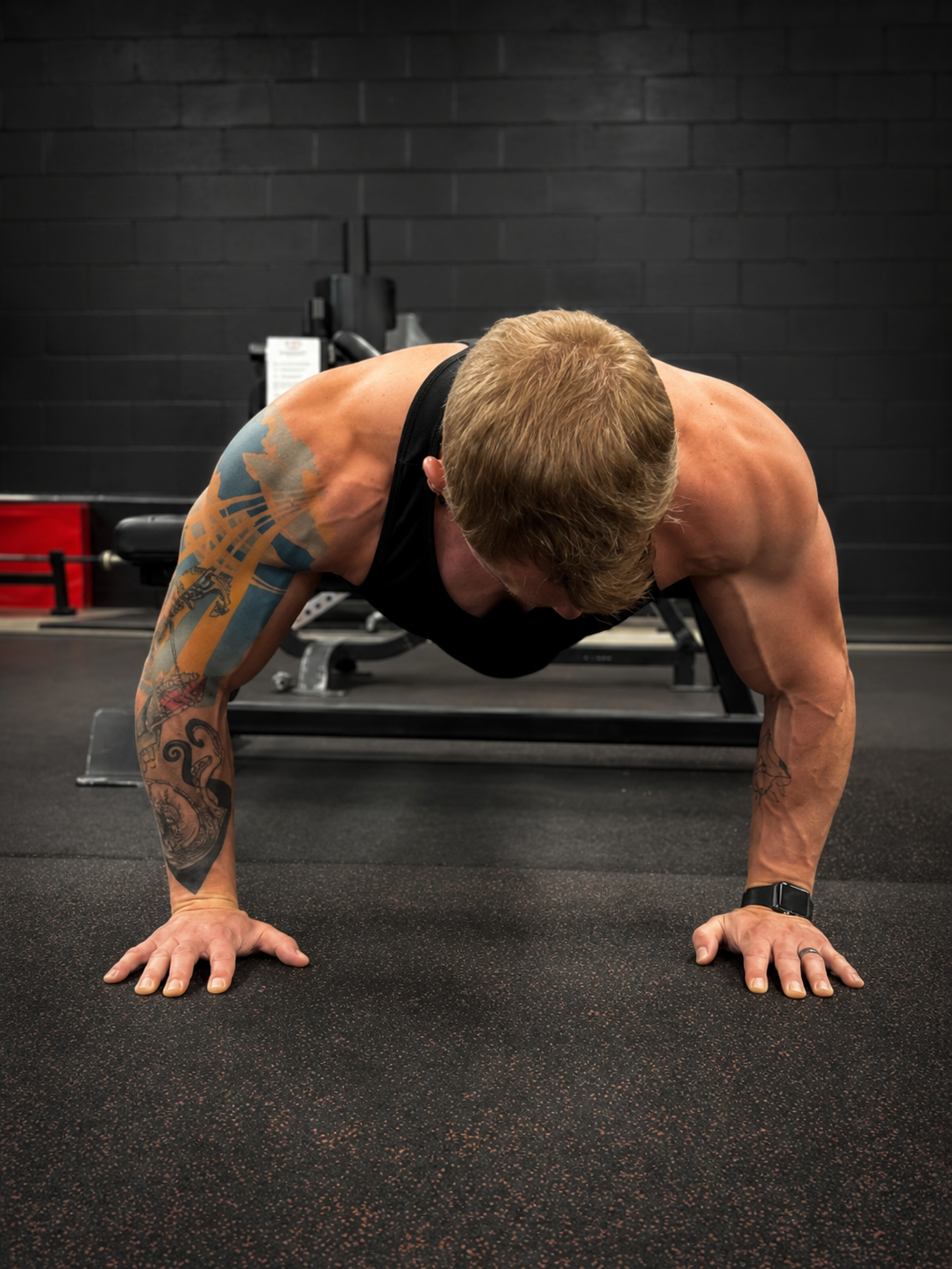 A man with tattoos on his arms is doing push-ups on a black gym floor, with a black brick wall and gym equipment in the background.