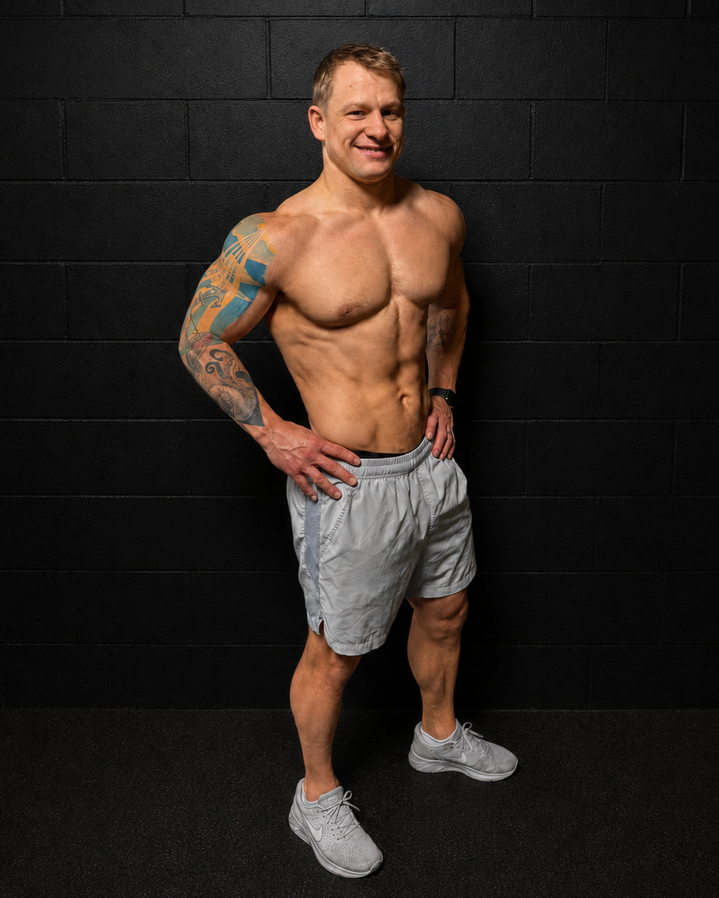 A muscular, shirtless man with tattooed arms, wearing gray shorts and white sneakers, standing against a black wall, smiling confidently.