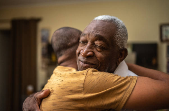 An older man with gray hair and a white shirt hugging a younger man wearing a yellow T-shirt in a warm embrace in a cozy indoor setting.