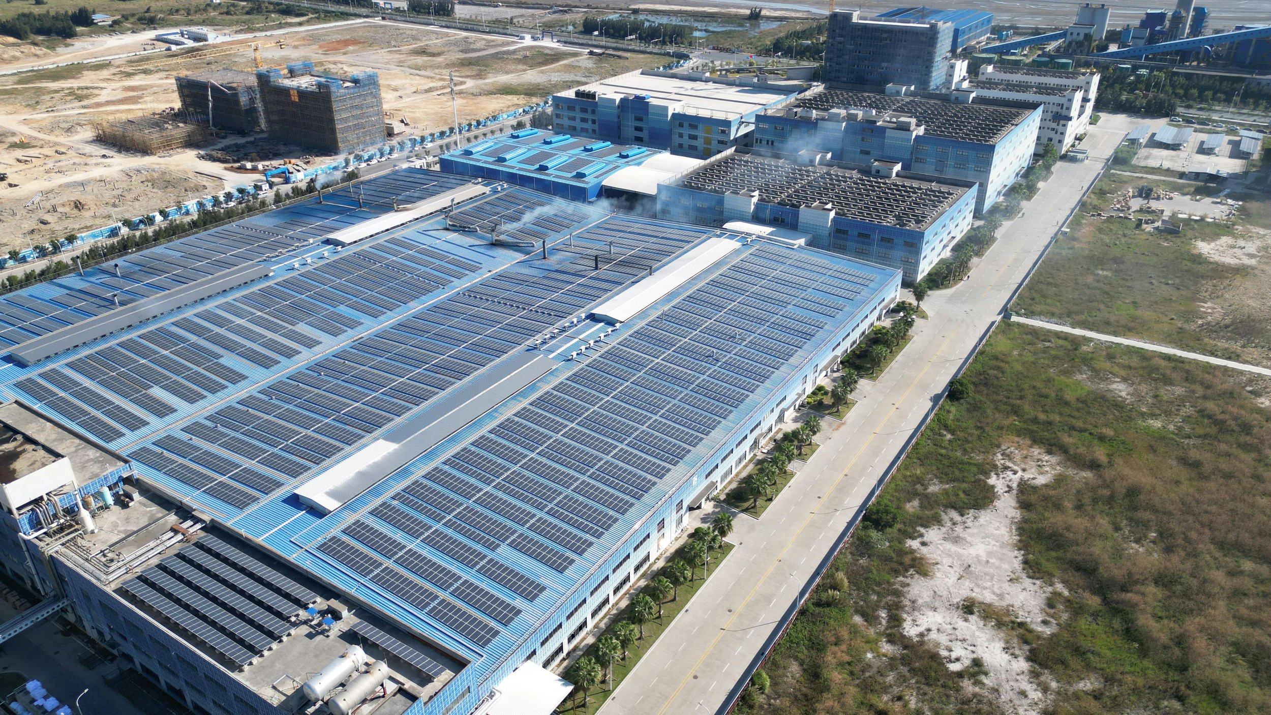 Aerial view of a large industrial building with a roof covered in solar panels, surrounded by roads, empty lots, and other buildings under construction.